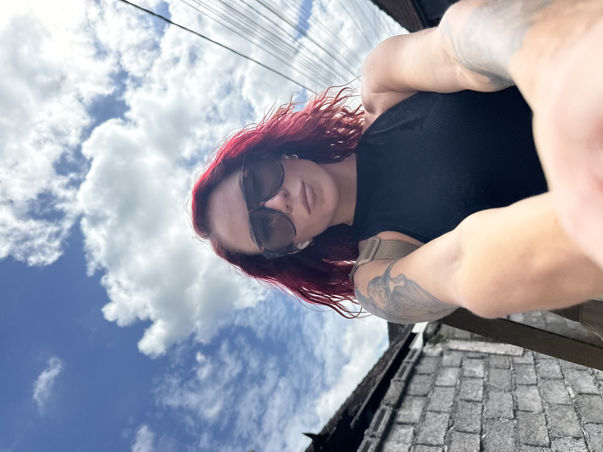 A low-angle, close-up selfie shows a light-skinned person with red hair and sunglasses. The person is wearing a black shirt and stands on a rooftop with gray tiles. A partially visible arm with a tattoo is in the foreground. The background features a blue sky with white clouds and power lines. The location is likely in North Kuta, Indonesia.