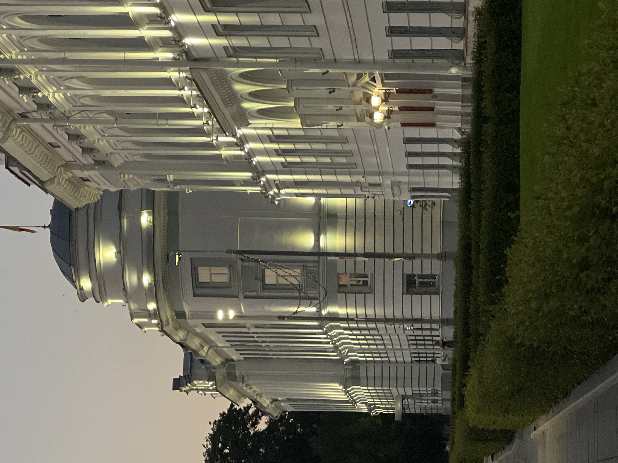 This image shows a stately white building with classical architectural elements, illuminated by warm yellow lights against a twilight sky. The building features a prominent dome on the left side, topped with a flagpole flying a yellow flag. The facade on the right is characterized by a series of arched windows and columns. In the foreground, a well-maintained green hedge lines a paved walkway. Two individuals are visible standing near the hedge, appearing to be conversing. The scene suggests a public space or administrative building in Tver, Russia, likely during the early evening hours.