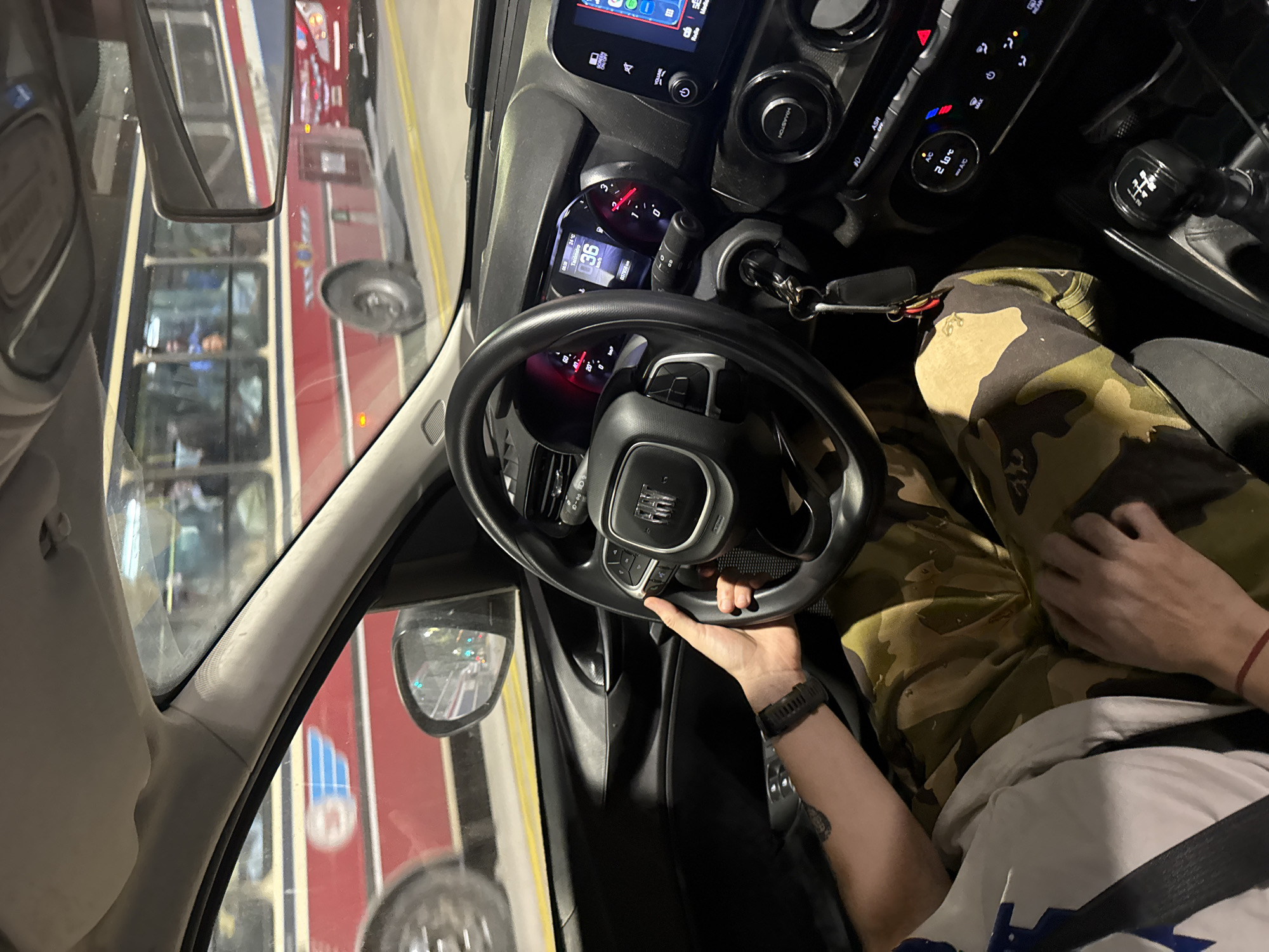 The image depicts the interior of a car with the driver visible in the lower right portion of the frame. The driver is wearing a white t-shirt, camo pants, and a smartwatch. The car's dashboard, steering wheel, and controls are visible, including the gear shift and the infotainment system. The car is stationary and the key is in the ignition. Through the car's window, a red bus is visible, indicative of public transportation. Based on the bus design (red and white color scheme with blue accents), it is likely a bus in the city of Buenos Aires, Argentina.