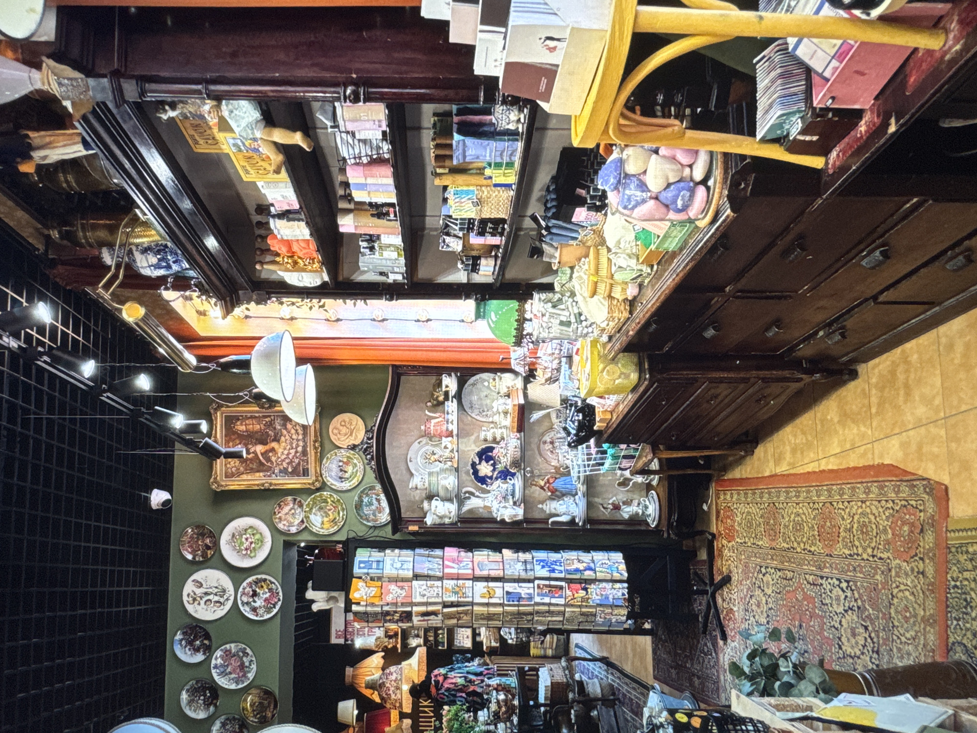 The image depicts an interior view of a shop, likely an antique or curio store, located in Novinki, Russia. The shop is filled with a variety of decorative and functional items. Dominating the right side of the frame is a tall, dark wooden cabinet with multiple shelves displaying an assortment of merchandise. These items include bottles, boxes, and decorative objects. Below the shelves, a dark wooden dresser with several drawers is visible, topped with more displayed items such as baskets, jars filled with colorful objects, and small boxes.
On the left side of the image, a green wall is adorned with numerous decorative plates. In the center, a tall rotating display rack filled with postcards or small booklets is positioned. Further to the left, antique lamps with patterned shades are visible, along with other display shelves and furniture. The floor is covered with Persian-style rugs. The overall atmosphere suggests a cluttered yet curated retail environment, common in shops selling vintage or artisanal goods. The lighting is a mix of overhead fixtures and display lighting, creating a warm and inviting ambiance. There are no visible people actively engaged in any action within the frame.