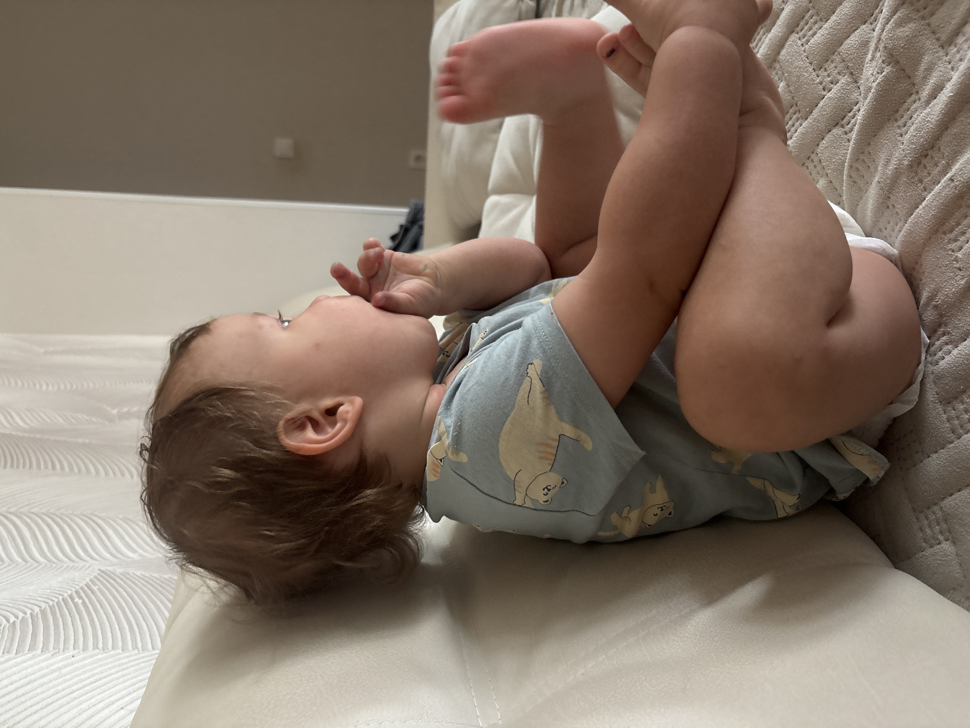 The image captures a baby lying on its back on a light-colored, textured surface, likely a couch or bed, in an indoor setting in Chaikovsky, Russia. The baby, an infant or young toddler with light brown hair, is dressed in a light blue short-sleeved top or onesie adorned with cartoon-like cat or bear figures.
The primary activity shows the baby with its legs raised and bent, bringing its feet up towards its chest. One of the baby's hands is positioned near its mouth or chin, suggesting self-soothing, playing, or exploration. The baby's gaze is directed slightly upwards and to the left, appearing engaged with something out of frame or simply contemplative.
Notable details include the comfortable, domestic environment. The surface on which the baby lies is a pale, possibly cream or off-white fabric, exhibiting a woven or knitted pattern on the right side of the frame. Another smooth, lighter surface is visible under the baby's left side and head. The background consists of a plain light wall or furniture item, topped by a darker, neutral-colored wall. The lighting is soft and consistent, indicating either daytime indoors or a well-lit interior. No text is visible in the image. The overall impression is one of a peaceful, everyday moment.