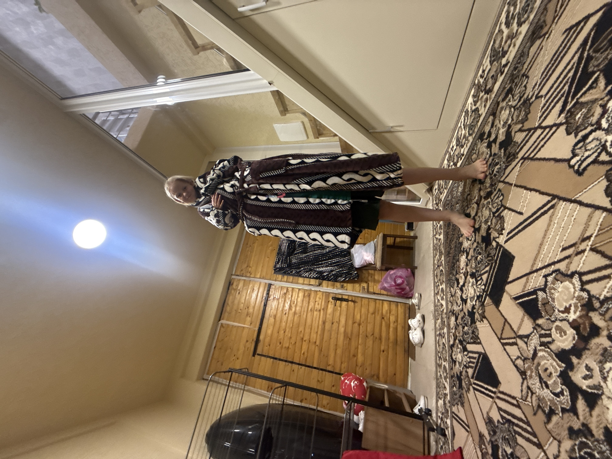A full-length medium shot shows a person indoors with a wooden wall and a rug nearby. The person is holding a dark brown robe and appears to be on a rug with a beige and brown geometric design. There are dark and light patches throughout the room, and a circular light shines brightly on the left side of the frame. There appears to be an open door near the top left side of the picture.