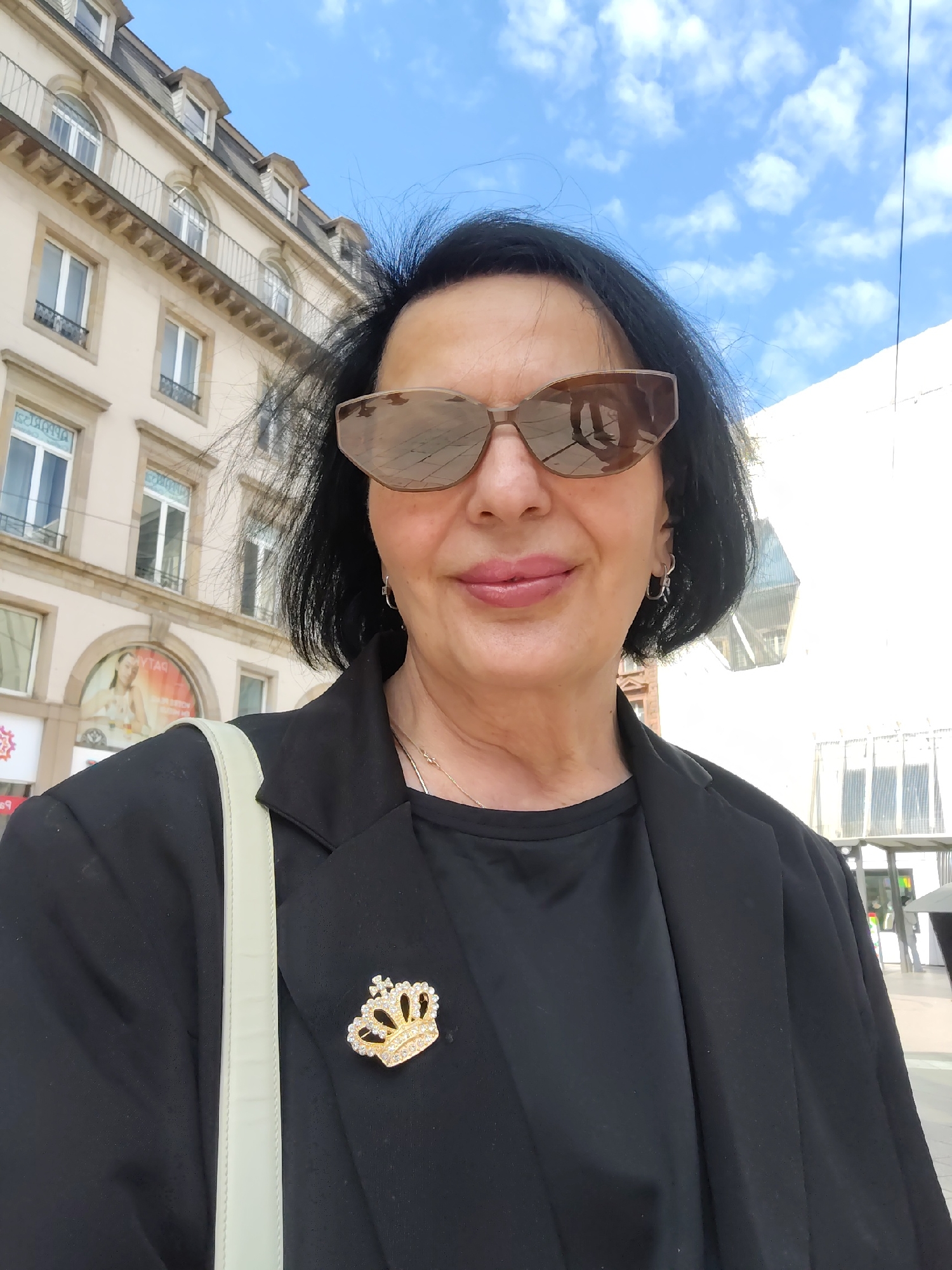 A woman wearing sunglasses, a black blazer with a crown-shaped brooch, and a light-colored shoulder bag stands on a city street. The building in the background has a row of arched windows and a sign with text in an unidentifiable language. This photograph was taken in Strasbourg, France.