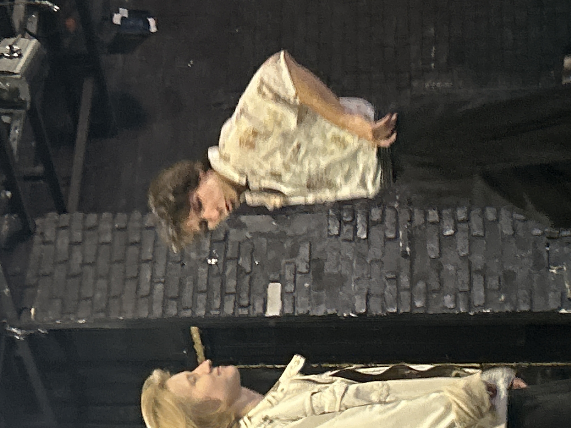 In an indoor setting with a dark, rough brick wall as the backdrop, two young individuals are visible. The person on the left, seen in profile, has light-colored hair and is wearing a light-colored jacket over a patterned shirt. They are looking towards the right. The person on the right is facing partially towards the left, with tousled brown hair. They are wearing a short-sleeved collared shirt with a floral or patterned design in muted tones of yellow and white, and dark pants. Their left hand is resting on their hip. The lighting suggests an artificial environment, possibly a stage or studio. The overall scene is dimly lit, with focus on the individuals and the textured brick wall.