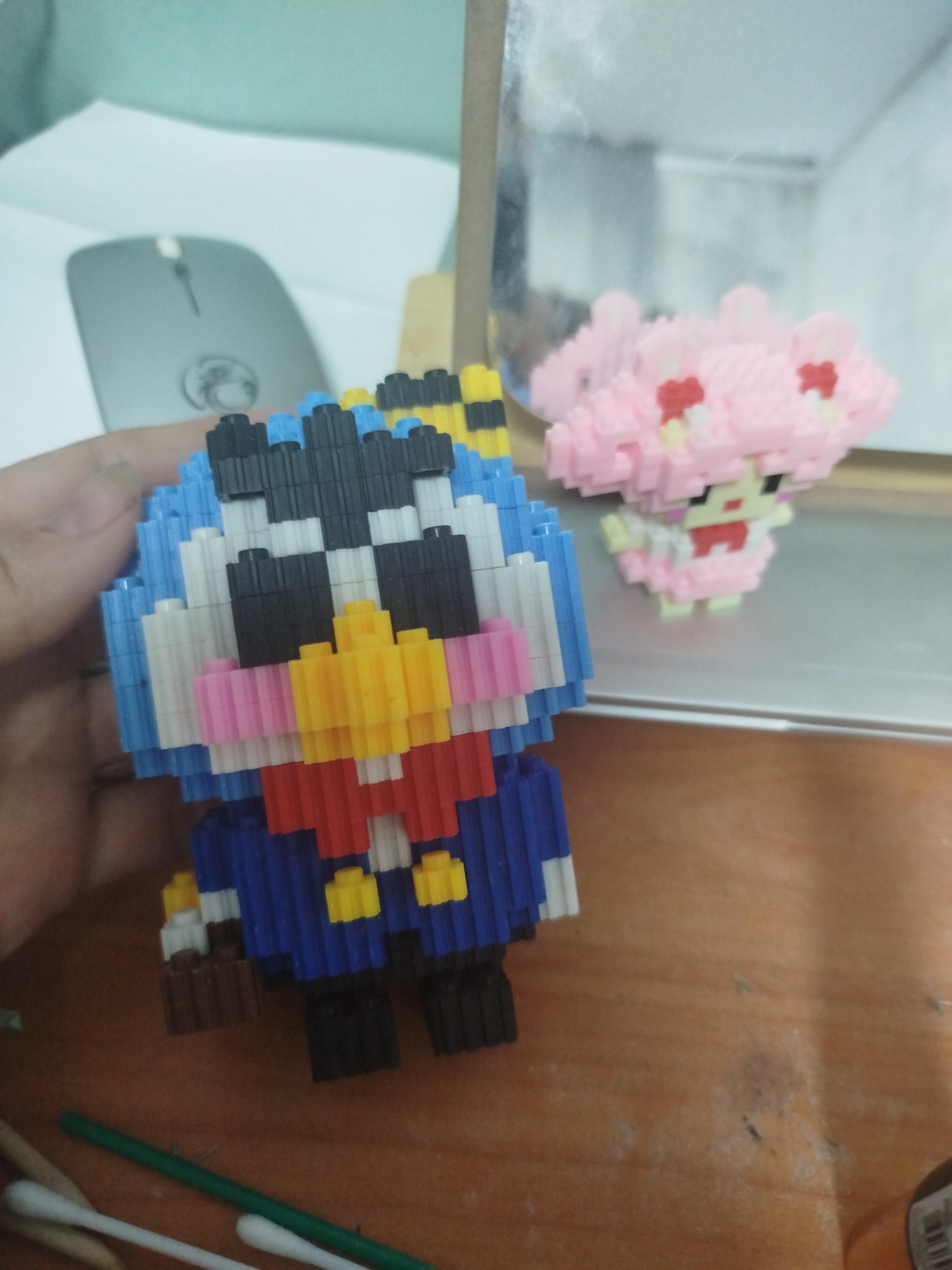 The image displays two pixelated toy figures on a wooden surface. The figure in the foreground, held by a person's hand, is blue, white, black, yellow, pink, and red, resembling a bird-like character with a suit and a bow tie. To the right and slightly behind, a pink pixelated figure with a dress and heart accents is visible, reflecting in a mirrored surface. In the background, a grey computer mouse is on a white surface. The scene is indoors, likely on a desk. The location of Zavodoukovsk, Russia, is not discernible from the image.