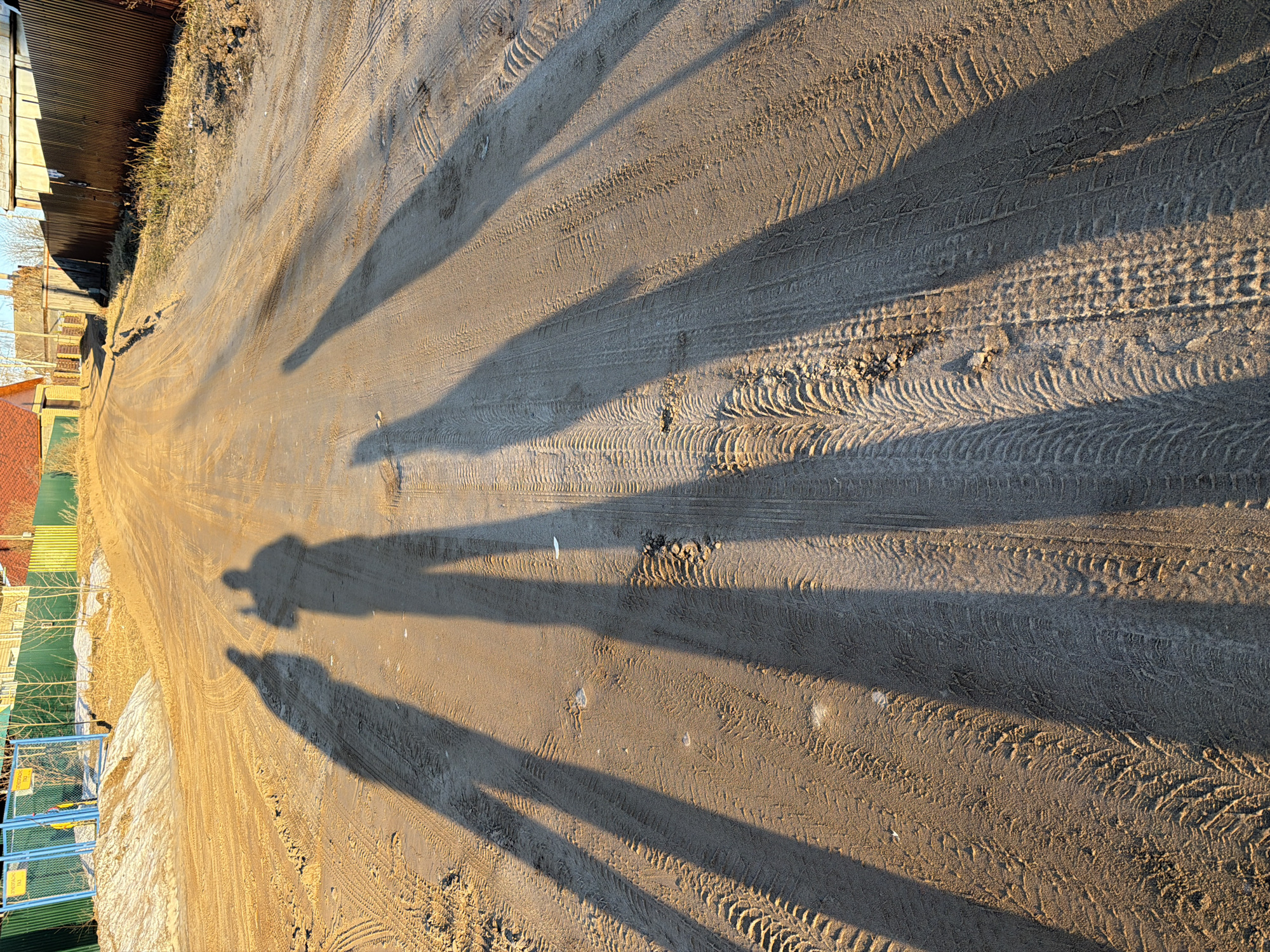 The image shows a dirt road in what appears to be a residential area in Chaikovsky, Russia. There are tire tracks running along the road's surface. The shadows of at least two people are visible, suggesting the presence of individuals taking the picture. A fence and buildings are on the road's edge. The dirt on the road surface indicates it is likely an unpaved road or alleyway. It appears to be a sunny day, based on the shadows and lighting. The road is a shade of light brown, with a fence to the left of the picture.