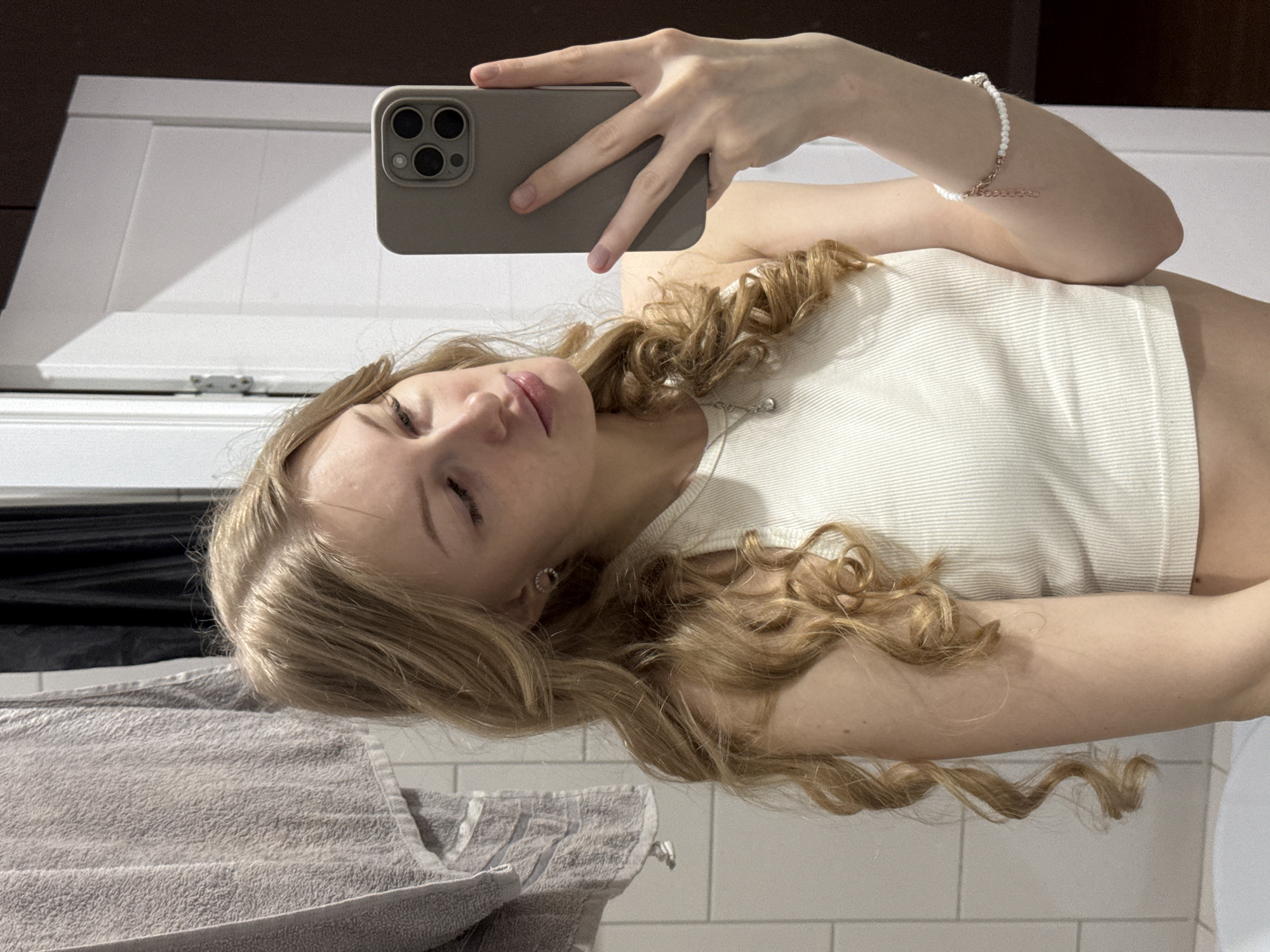 A young woman is taking a selfie in a bathroom. She is fair-skinned with blonde, curly hair. She wears a white tank top and a bracelet on her right wrist. She is holding a phone with a grey case. A grey towel is visible in the foreground. The background shows white bathroom tiles and a white cabinet. There is no indication of Perm, Russia in the image.