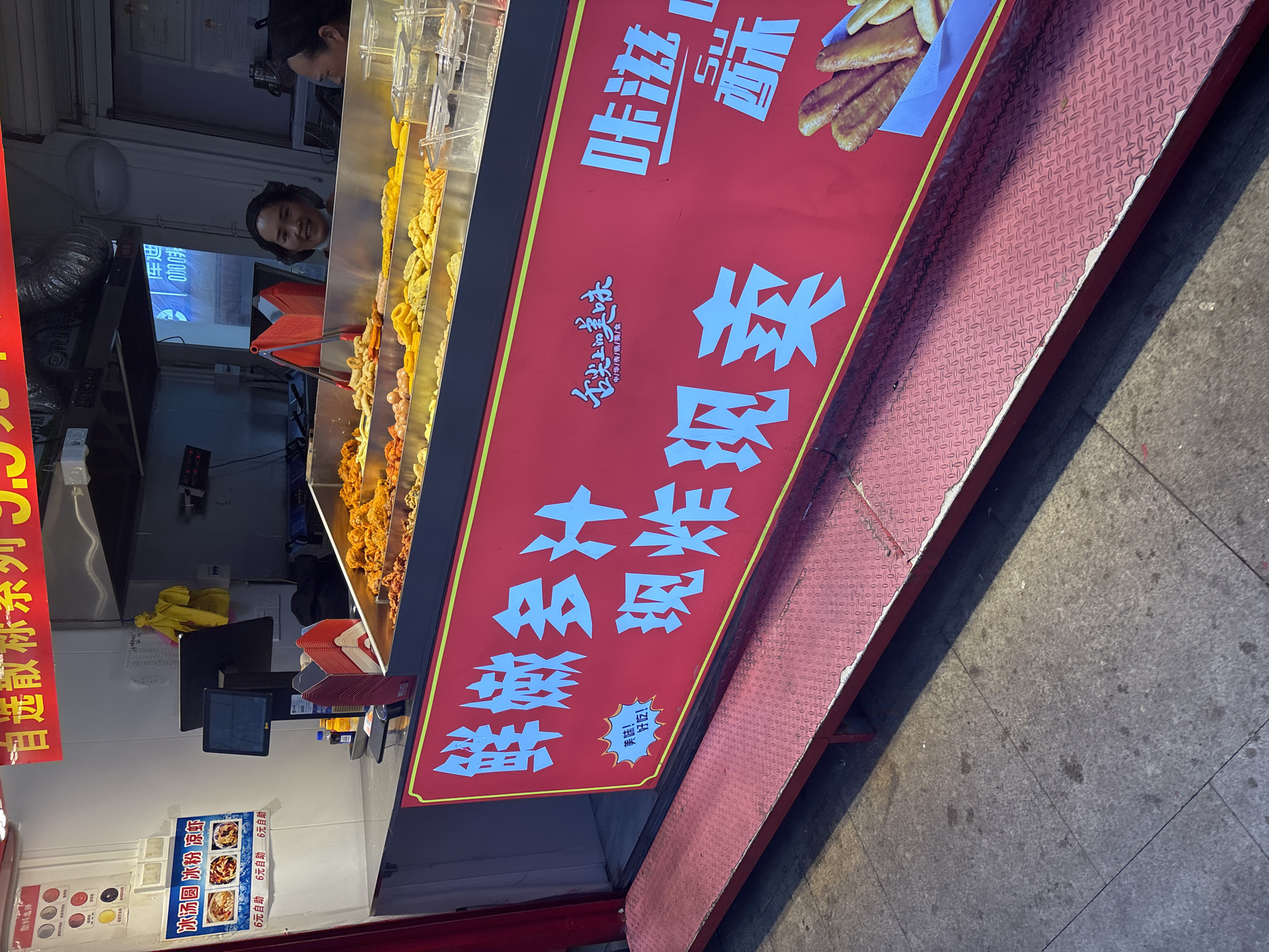 The image shows an outdoor food stall in Yongchuan, China, featuring a red sign in Chinese with white text, offering deep-fried food. A person is behind the stall looking into the camera, there is food on skewers in a glass display. There are also images and text promoting ice-cold desserts.