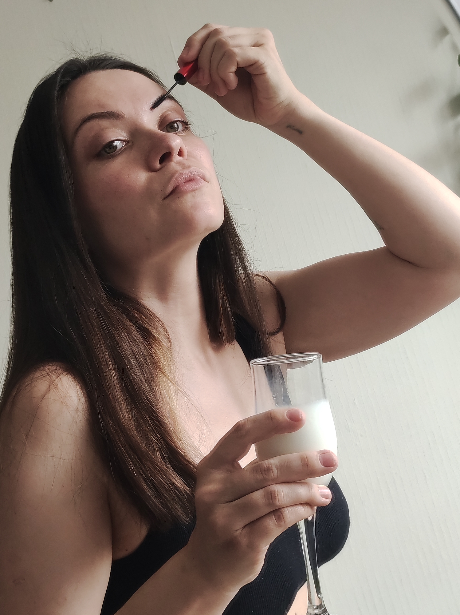A young woman is pictured from the chest up, holding a glass of milk in her left hand and applying makeup to her left eyebrow with her right hand. She is wearing a black top. Her dark brown hair is parted on the left and falls down her shoulder. She has a tattoo on her left forearm. The background is a plain, light-colored wall. There is no indication of the city of Chaikovsky, Russia in the image.