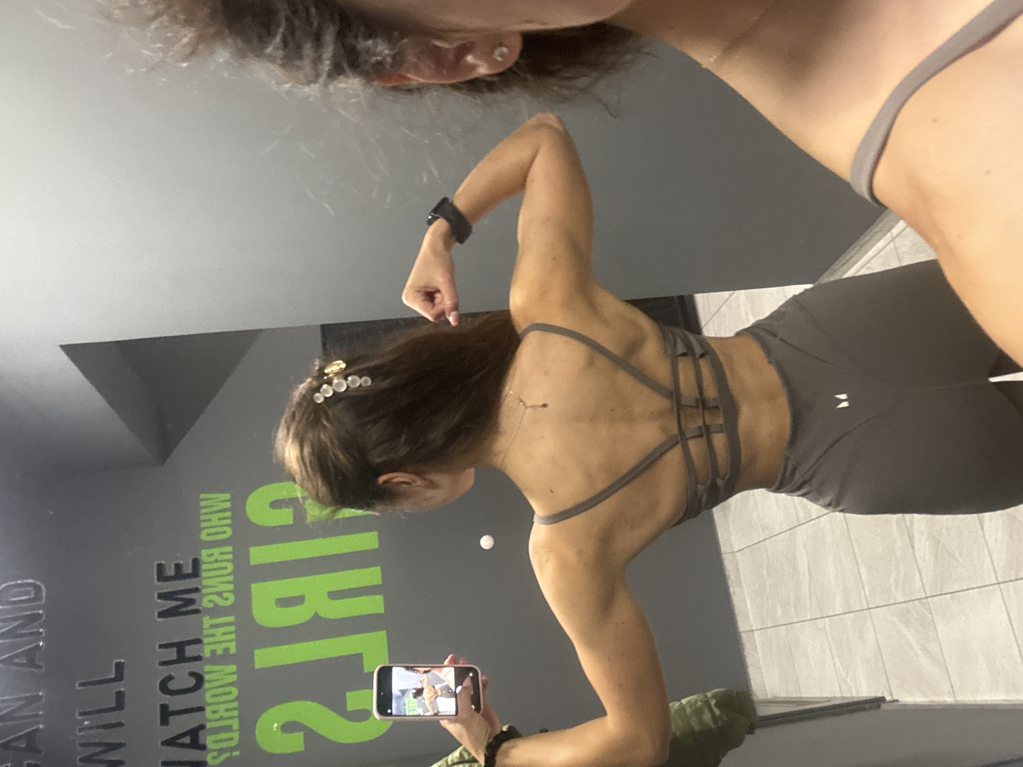 The image shows a young woman in gym attire taking a mirror selfie in a gym. The woman is facing away from the camera, with her back to the viewer, and is flexing her arm. She is wearing a gray sports bra and matching leggings. In the background, there is a mirror with text that appears to be motivational quotes. The text on the mirror reads, "GAIN AND WILL WATCH ME GIRLS". To the right of the woman, a portion of another person's face is visible in the mirror, also taking a selfie. The gym floor is tiled, and a portion of a locker or wall is visible on the left side of the image. The scene takes place indoors, in what appears to be a public gym facility. No specific location in Varna, Bulgaria is indicated in the image.