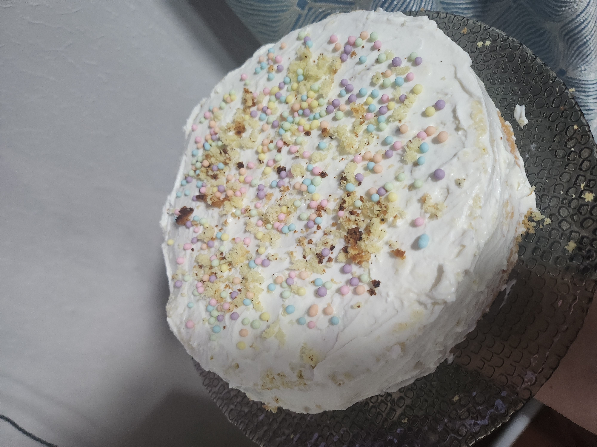 A frosted cake sits atop a textured glass plate. The frosting is white and unevenly applied. The top of the cake is decorated with small, pastel-colored spherical sprinkles and what appears to be cake crumbs. The cake is positioned against a light-colored background, with a partial view of a blue and white patterned curtain in the upper right corner. The cake is likely located in Krasnogorskoye, Russia.