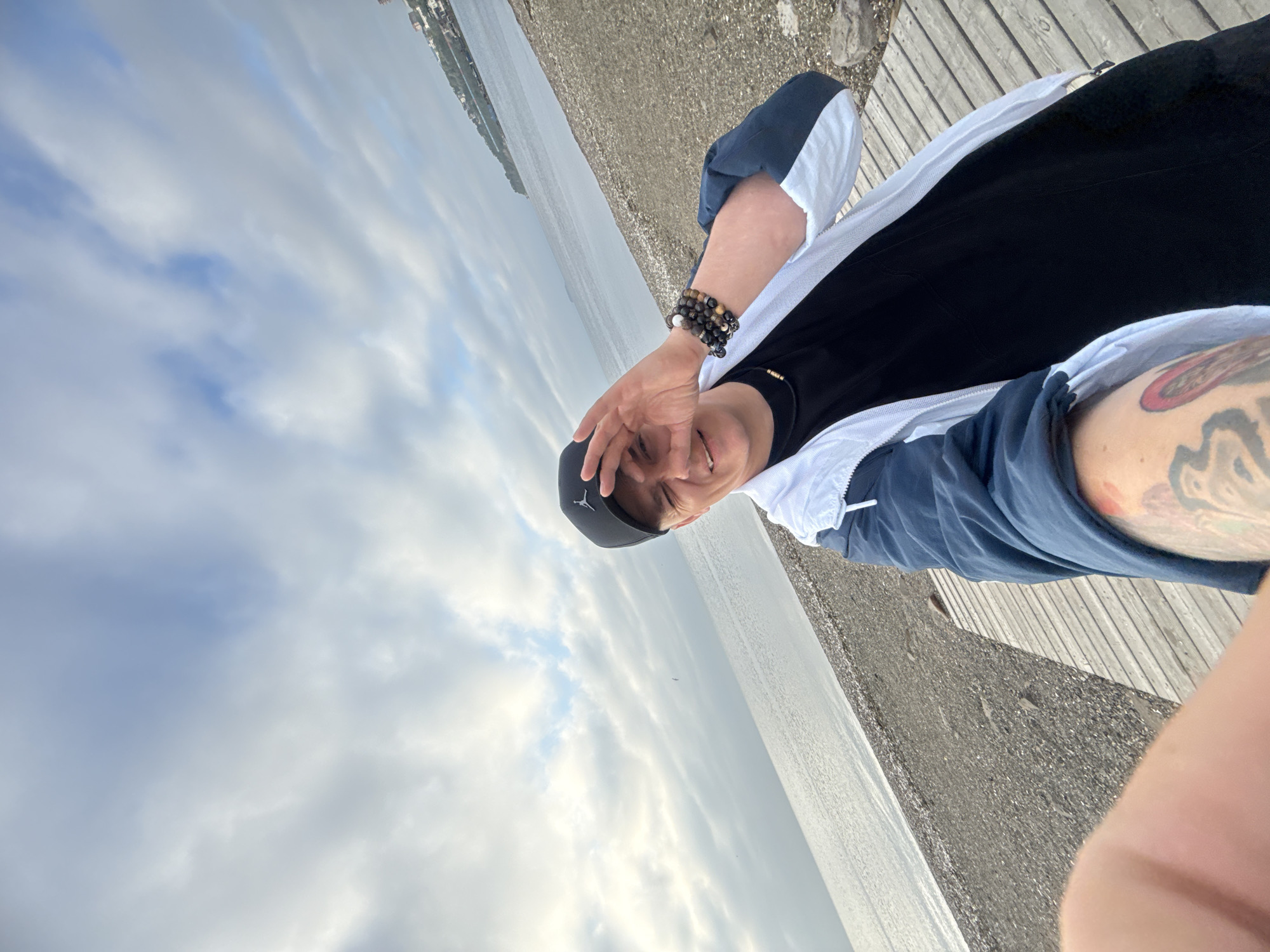 A young man wearing a black baseball cap with a Jordan logo, a black t-shirt, a blue and white jacket, and dark pants is taking a selfie on a beach in Vladivostok, Russia. He is wearing bracelets on his right wrist. The man is smiling and has his right hand covering his eye. The beach has a gravelly surface and a wooden boardwalk. The sea is calm and the sky is cloudy. In the background, there are some buildings on a hill and a body of water.