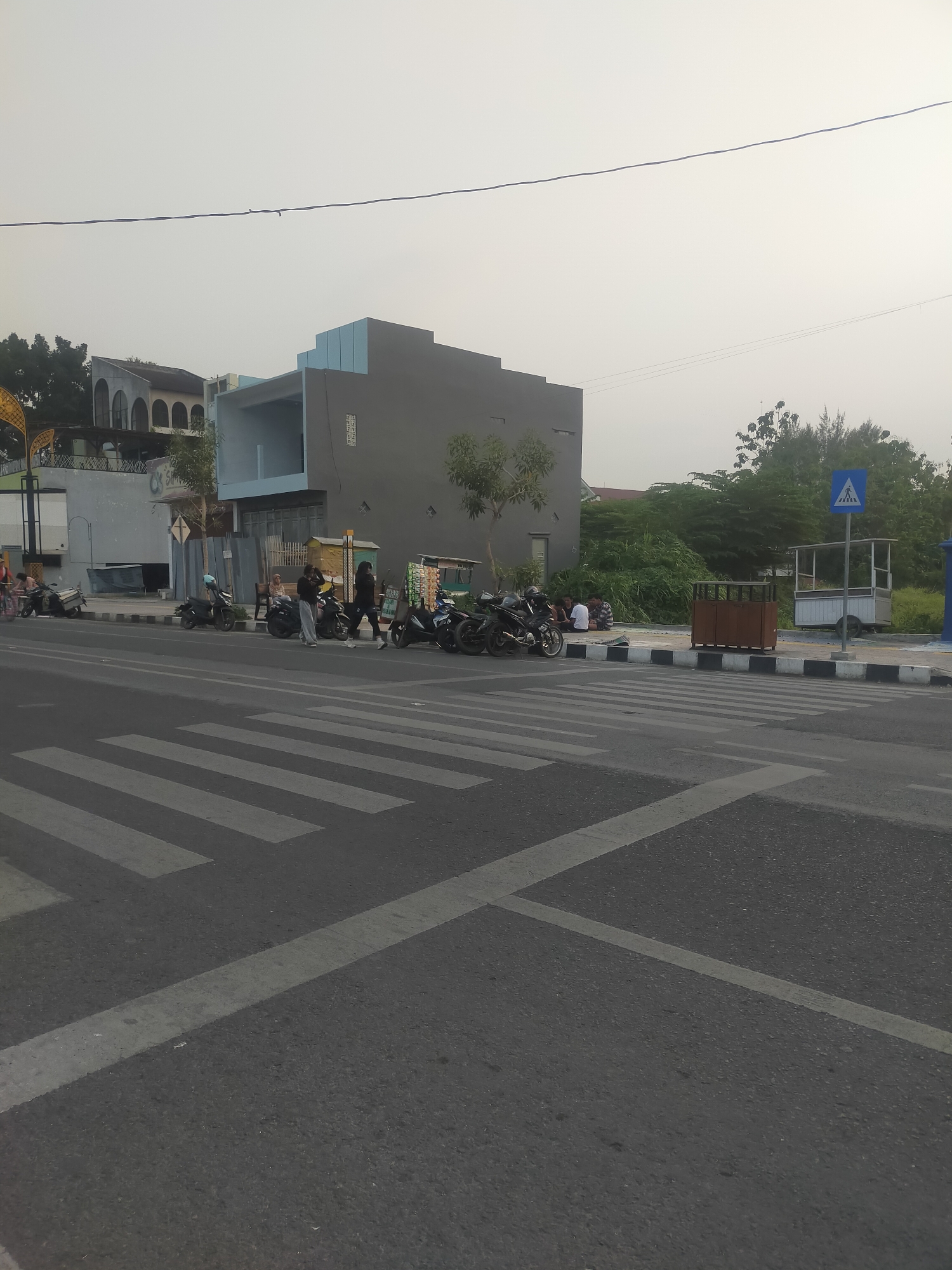 The image shows a street scene in Slawi, Indonesia, with a pedestrian crossing in the foreground. Several motorcycles are parked along the sidewalk, and a group of people are gathered near them. In the background, there is a building with a modern, angular design, and some trees and vegetation. A pedestrian crossing sign is visible on the right side of the image. The sky is overcast, and the overall lighting suggests it is either early morning or late afternoon. The scene appears to be a typical urban environment with moderate activity.