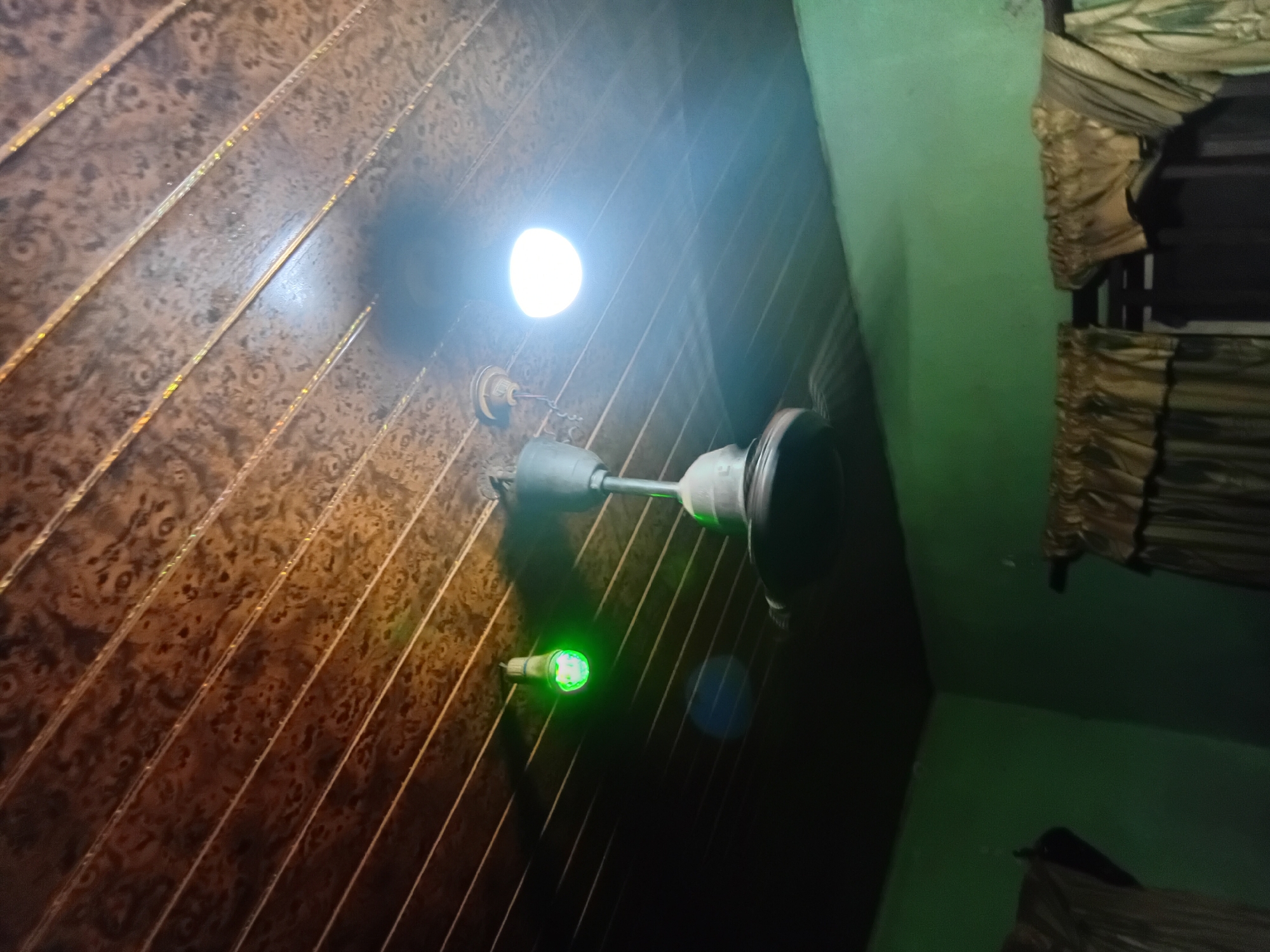 The image shows an interior view of a room in Nkpor, Nigeria, likely a bedroom or living area. A ceiling fan is suspended from a wooden, patterned ceiling. To the left of the fan, a green LED light bulb is illuminated, casting a greenish glow. To the right of the fan, a bright white light bulb is also illuminated. The walls of the room are painted a light green. Curtains, appearing to be made of a golden or beige fabric with some sheen, hang in the background, partially obscuring windows or doorways. The lighting conditions are mixed, with the stark contrast between the green and white bulbs creating an unusual ambiance. The scene appears to be captured from a low angle, looking upwards towards the ceiling.