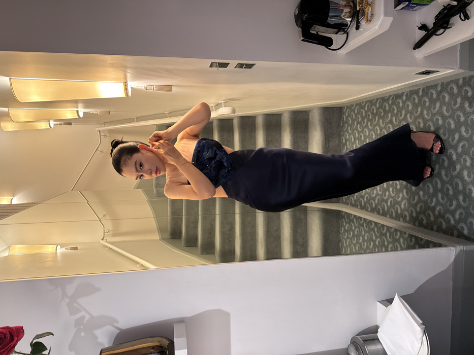 A woman in a strapless navy gown poses near a staircase with gray carpet and a white railing. The dress features a floral detail on the bodice and a slim fit. She is wearing her hair in a bun and has a delicate bracelet on her wrist. The staircase is illuminated by wall-mounted lights. Other items, including a hairdryer, hair straightener, and various toiletries, sit on a shelf. A vase of red roses is visible at the bottom of the frame.