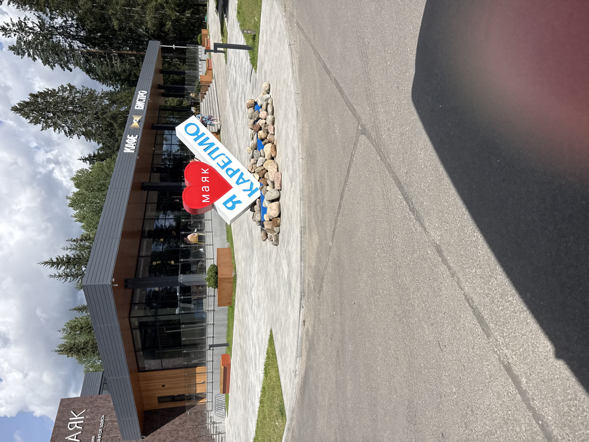 The image depicts a modern building with a dark gray roof and large glass windows, housing a "КАФЕ" and "БИСТРО." In front of the building, a large, stylized red heart with the word "МАЯК" (Lighthouse) written on it is positioned next to a white sign that reads "Я КАРЕЛИЮ" (I Karelia). This monument is surrounded by a collection of natural stones. The building is situated on a paved area with stone tiles and grass patches. Several trees are visible in the background, suggesting a location surrounded by nature, possibly within the city of Yakkima, Russia, given the context of the monument. The sky is cloudy with patches of blue. A shadow of a person or object falls across the foreground asphalt.