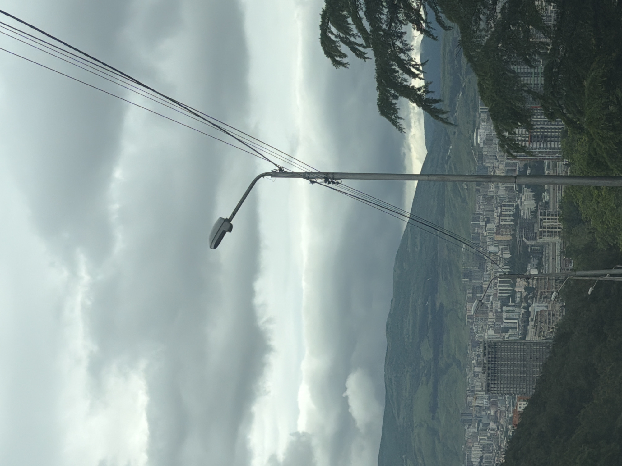 A street lamp stands prominently against a backdrop of a cloudy sky over Tbilisi, Georgia. A mountain range and a densely populated urban area fill the midground. The foreground is framed by a leafy tree. Power lines intersect the view.