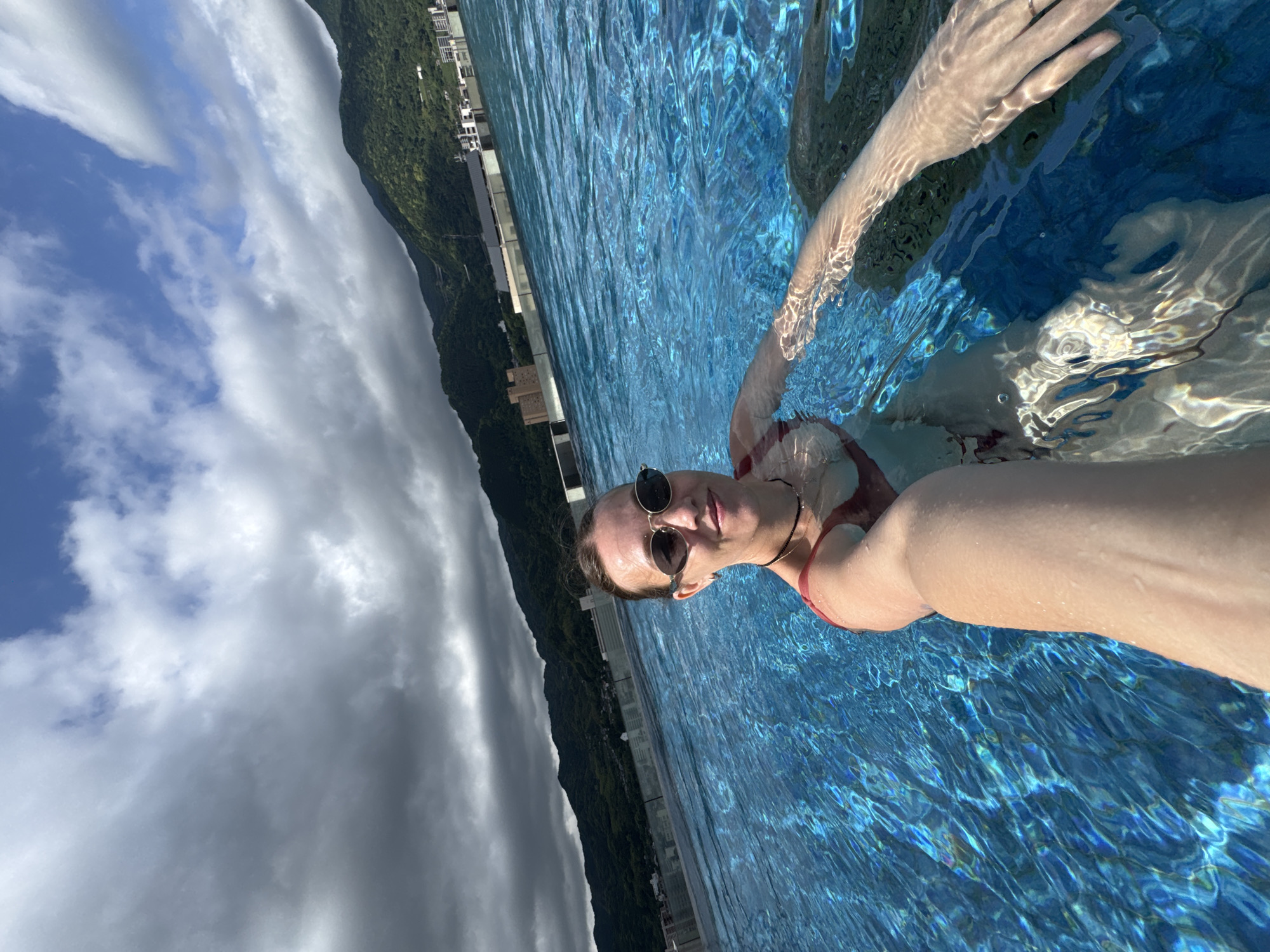 A woman wearing sunglasses and a red swimsuit is partially submerged in a blue-tiled swimming pool. The pool is an infinity pool with a view of a green, mountainous landscape and buildings beyond the pool's edge. Above the landscape, the sky is visible with a mix of blue sky and white clouds. The image appears to have been taken at a tilted angle. This appears to be an infinity pool in Beppu, Japan.
