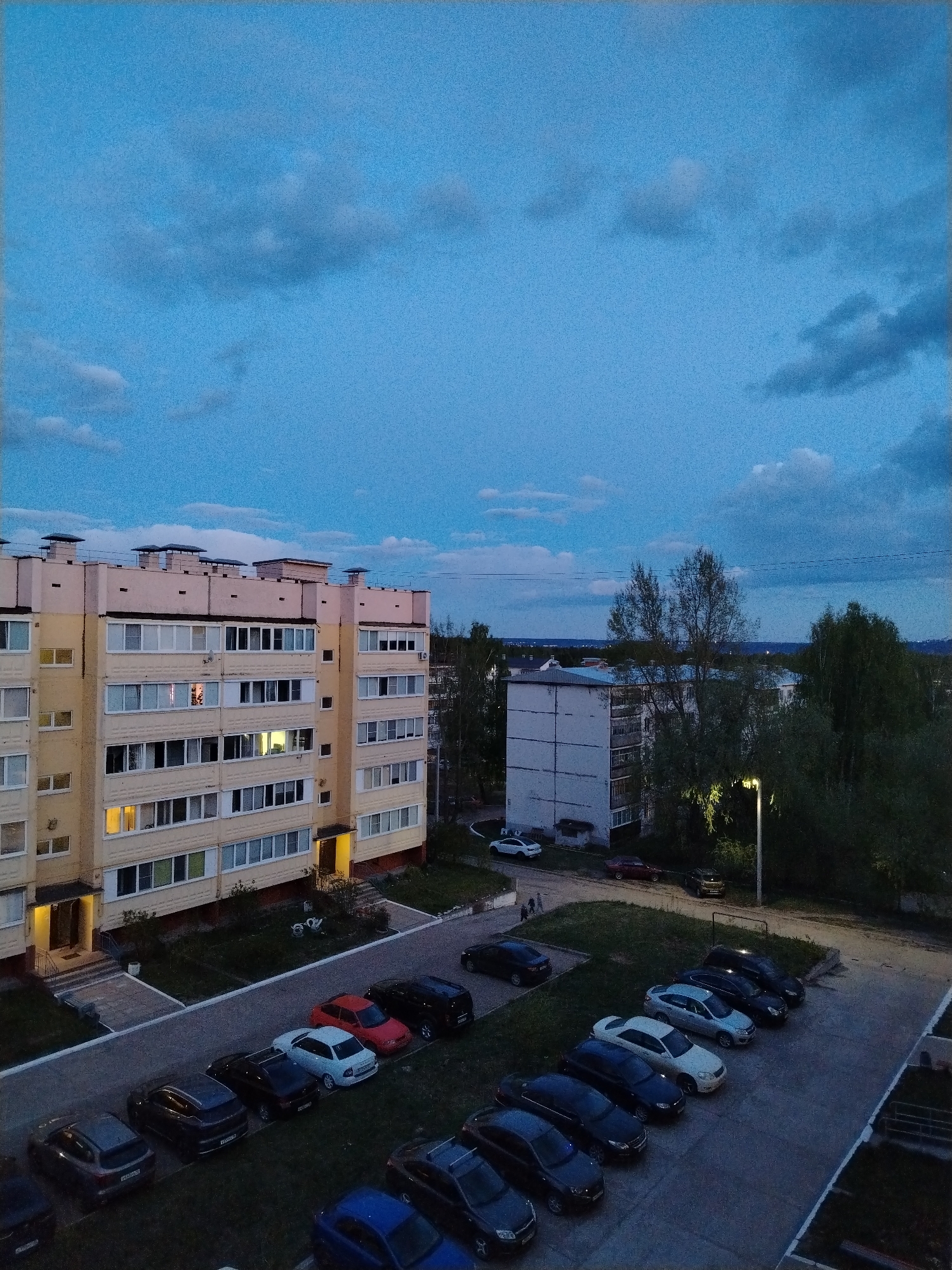 The image depicts an urban residential scene in Noviy Urengoy, Russia, at dusk. In the foreground, several rows of parked cars of varying colors and models fill a paved parking area. Adjacent to the parking area is a multi-story, light yellow apartment building with multiple windows, some illuminated, indicating occupancy. A secondary, smaller white building is visible behind the first building, partially obscured by trees. A streetlight illuminates the space between the buildings. Above, the sky is blue with scattered clouds.