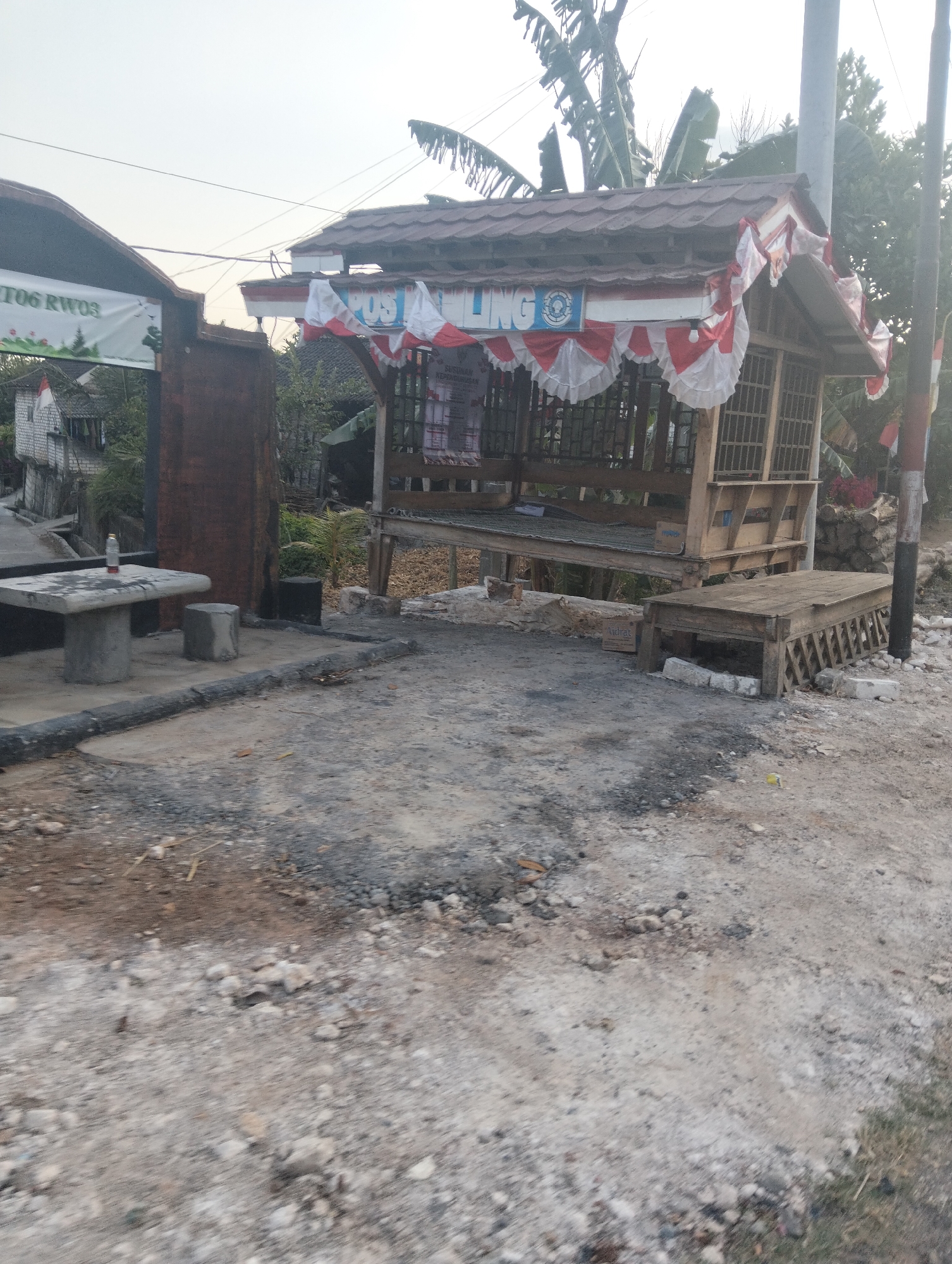 The image depicts a street scene in Dagan, Indonesia, with a focus on a small wooden kiosk decorated with red and white fabric. The kiosk appears to be a roadside stall or shelter, with a tiled roof and open sides. A sign above the stall reads "POS LING," and a small bottle of water rests on a concrete table to the left. The ground is paved with gravel and dirt, indicative of a rural or developing area. In the background, there are glimpses of houses, trees, and a utility pole. The overall impression is that of a simple, utilitarian structure in a humble setting.