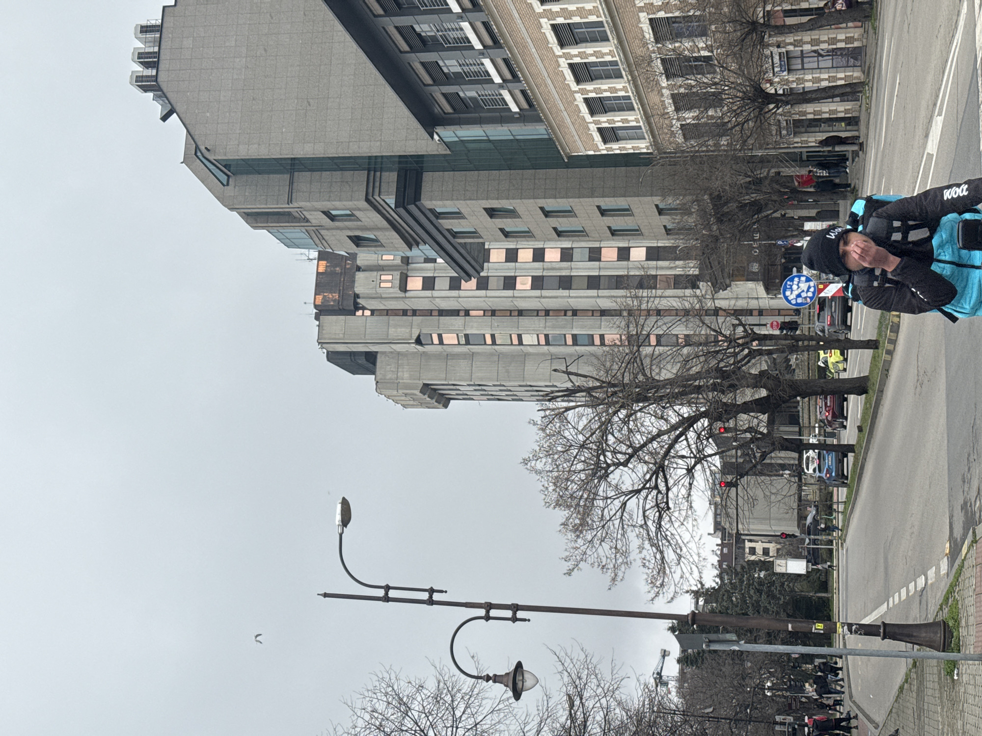 The image depicts an urban scene in Varna, Bulgaria. Dominating the left side of the frame are tall, grey, multi-story buildings with varying architectural styles, suggesting a mix of old and modern construction. A series of bare trees are scattered throughout the scene, indicating it may be autumn or winter. A vintage street lamp with two decorative lamp heads stands prominently in the foreground.
A delivery person wearing a black beanie, black jacket, and a bright blue delivery backpack is standing on the right side of the image, near the roadway. Cars and traffic signals are visible, suggesting an active street. Pedestrians can be seen walking on the sidewalk.
The overall environment suggests a typical city street in Varna, with a blend of buildings, infrastructure, and human activity. The sky appears overcast.