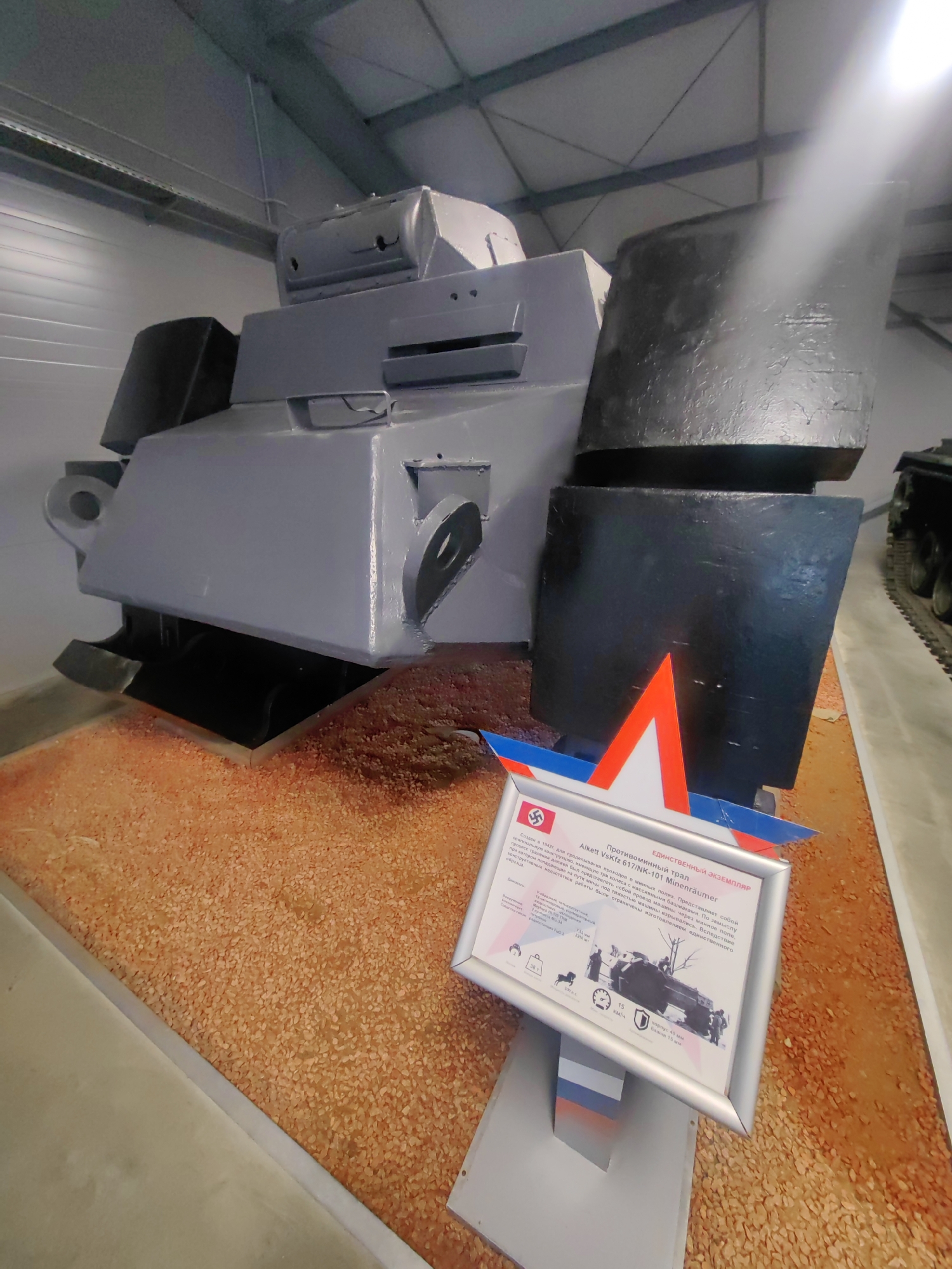 The image shows a museum exhibit featuring a German-designed armored vehicle, identified as an Alkett VkKfz 617/NK-101 Minenräumer. This vehicle is gray and black, with a large, angular structure. It is displayed on a textured orange-brown substrate, resembling small pebbles or gravel. In the foreground, a silver-framed informational display stands at an angle. The display contains text in Russian and German, along with an illustration of the vehicle and technical specifications. A red and white geometric decal is affixed to the side of the vehicle, just above the display. The exhibit is located within a large indoor space with a white paneled ceiling and visible structural beams, characteristic of a museum hall. The overall scene is well-lit, with a bright light source visible in the upper right corner. The location is identified as the Patriot Congress and Exhibition Center in Kubinka, Russia.