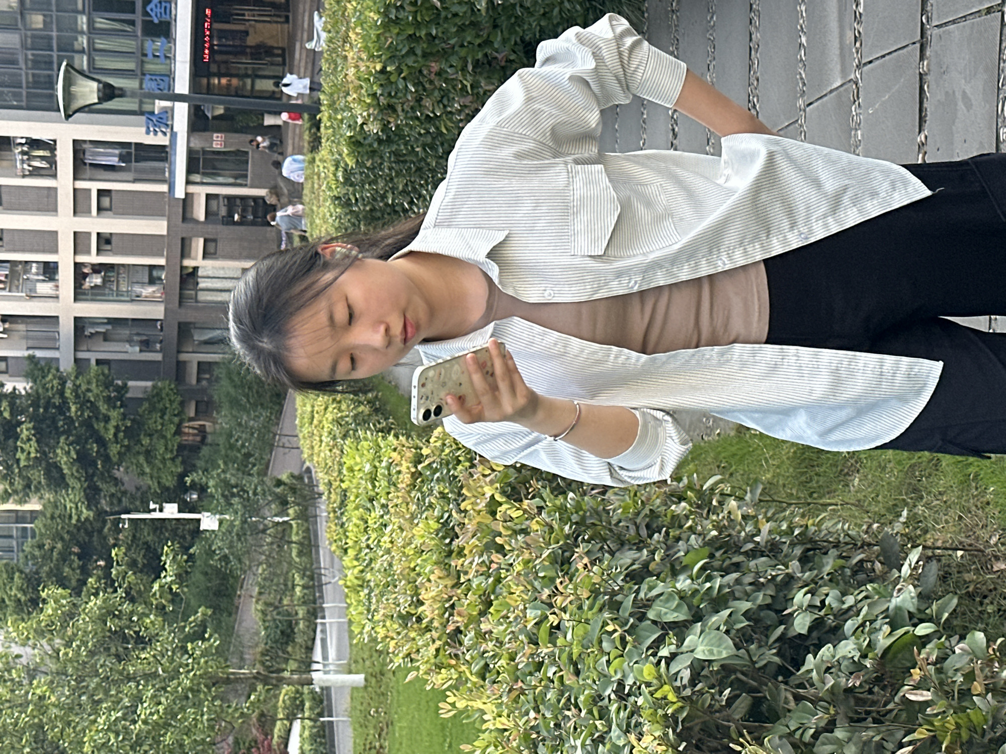 A young woman is standing outdoors in Jiangjin, China, looking at a phone. She is wearing a light-colored, striped, button-down shirt over a brown top and black pants. The background includes buildings, trees, and manicured bushes. The ground appears to be a combination of pavement and grass.