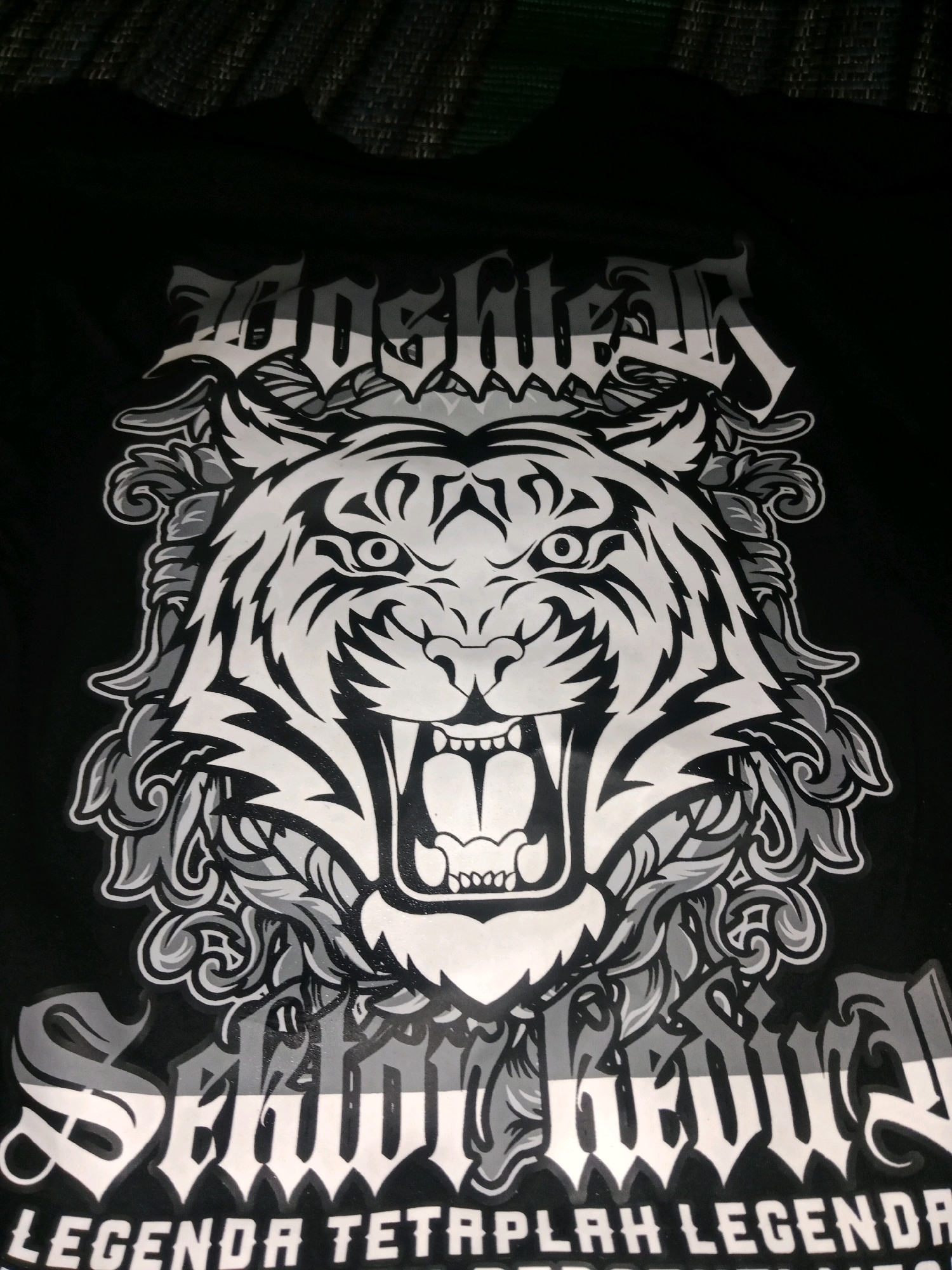 This image shows a black t-shirt with a graphic print. The graphic features a stylized, fierce tiger head in black and white, surrounded by ornate, swirling patterns. Below the tiger, there is text in a gothic font, with the words "SEMANGAT JUANG" clearly visible. At the very bottom of the image, there is more text which reads "LEGENDA TETAPLAH LEGENDA". The t-shirt is likely part of a collection or brand associated with the city of Ringinrejo, Indonesia, given the textual elements present. The context suggests a display or advertisement of this merchandise, possibly in a retail setting or promotional material originating from that location.