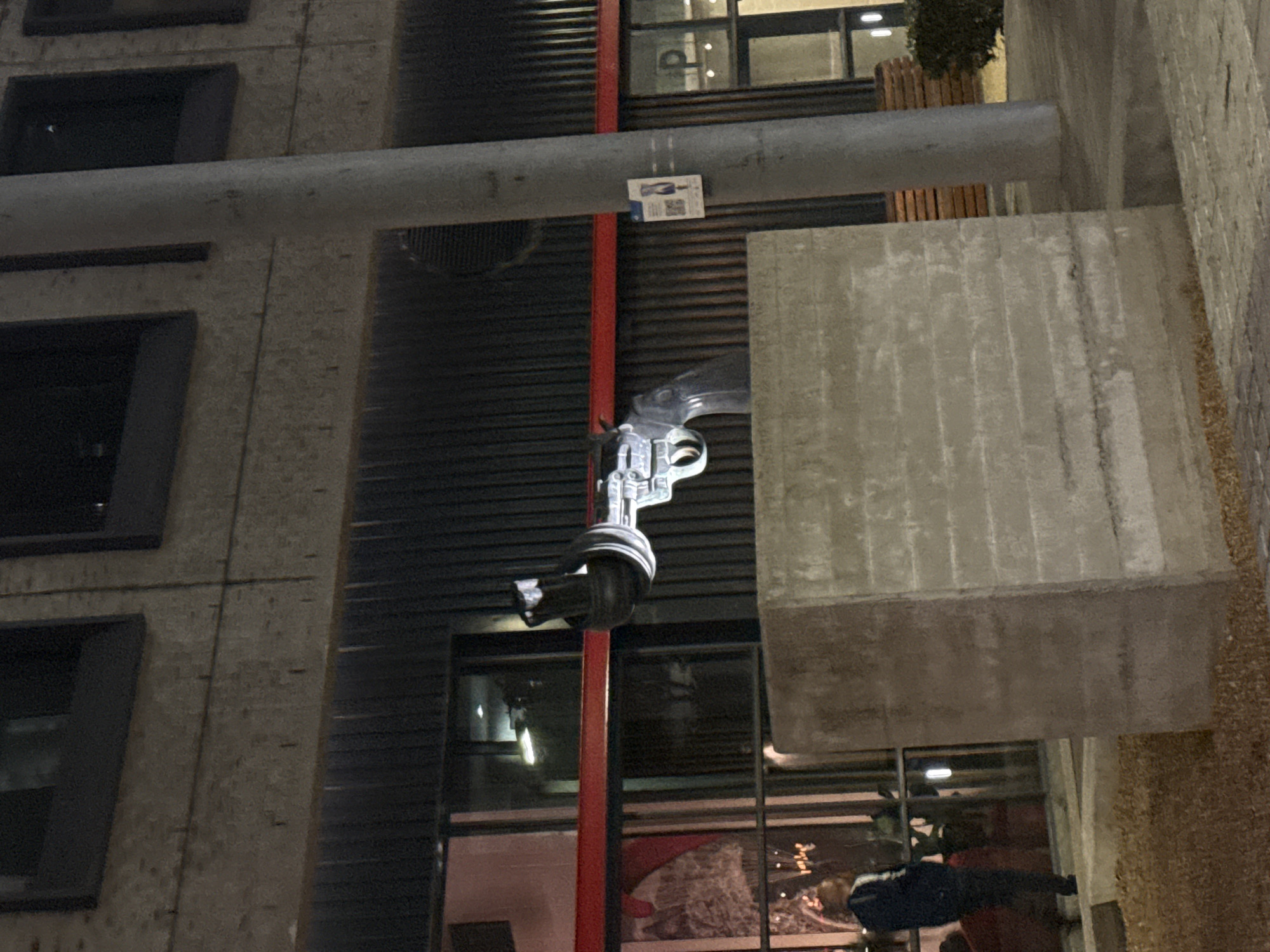 The image captures a nighttime urban scene at the V&A Waterfront in Cape Town, South Africa. The central subject is a sculpture, known as "Non-Violence," depicting an oversized silver-grey revolver with a dark handle and its barrel tied in a knot. This sculpture rests on a textured grey concrete rectangular plinth positioned slightly off-center to the left.
Behind the sculpture, a multi-story building features a facade composed of dark corrugated metal panels and grey concrete blocks. Large dark-framed windows on the ground floor reflect interior lights and are bisected by a prominent red horizontal beam. A window on the far right displays a white capital letter "P".
To the far left of the frame, a male figure is partially visible, wearing a dark jacket with white stripes on the sleeves, standing near the building's windows, facing slightly away from the camera. A grey cylindrical utility pole stands to the right of the sculpture, with a white flyer featuring a QR code and a graphic of a person attached to it. A wooden bench and green shrubbery are discernible behind the pole. The ground in the immediate foreground consists of light-colored gravel, transitioning to paved sections further to the right. No direct interactions between individuals or objects are observable.