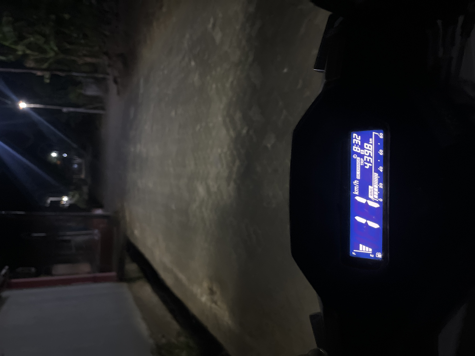 This is a night-time, point-of-view shot from a motorcycle in Karangrandu, Indonesia. The foreground features a digital motorcycle dashboard displaying time (8:32), trip distance (439.8 km), and fuel level. The road ahead is illuminated by streetlights and motorcycle headlights, revealing a paved road lined with houses and vegetation. The atmosphere is dark and quiet, typical of a rural Indonesian setting at night.