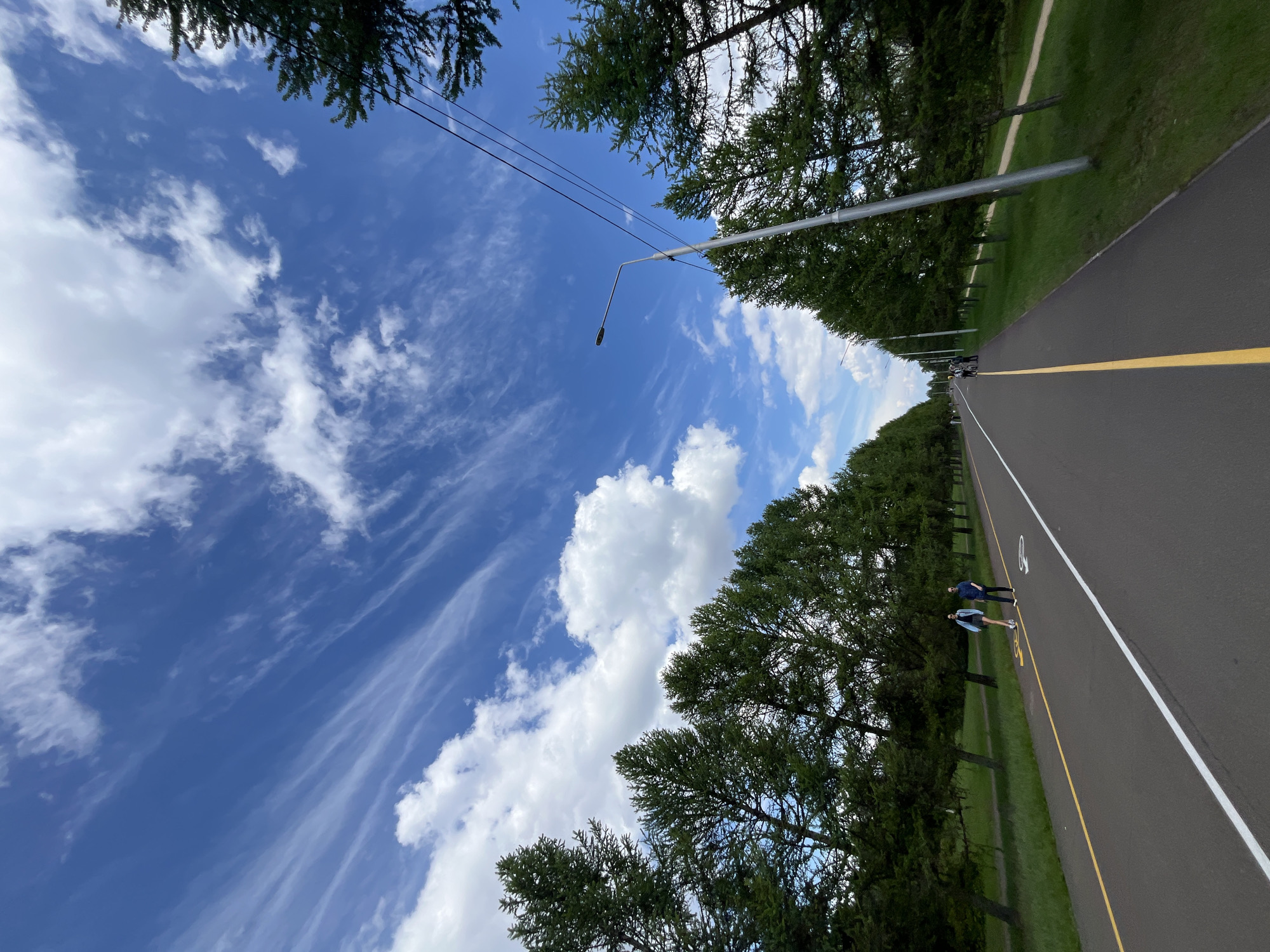 The image depicts a paved path in Krasnoyarsk, Russia, flanked by green grass and trees, under a partly cloudy blue sky. Two people are walking on the path. Streetlights line the path. A yellow line divides the path.