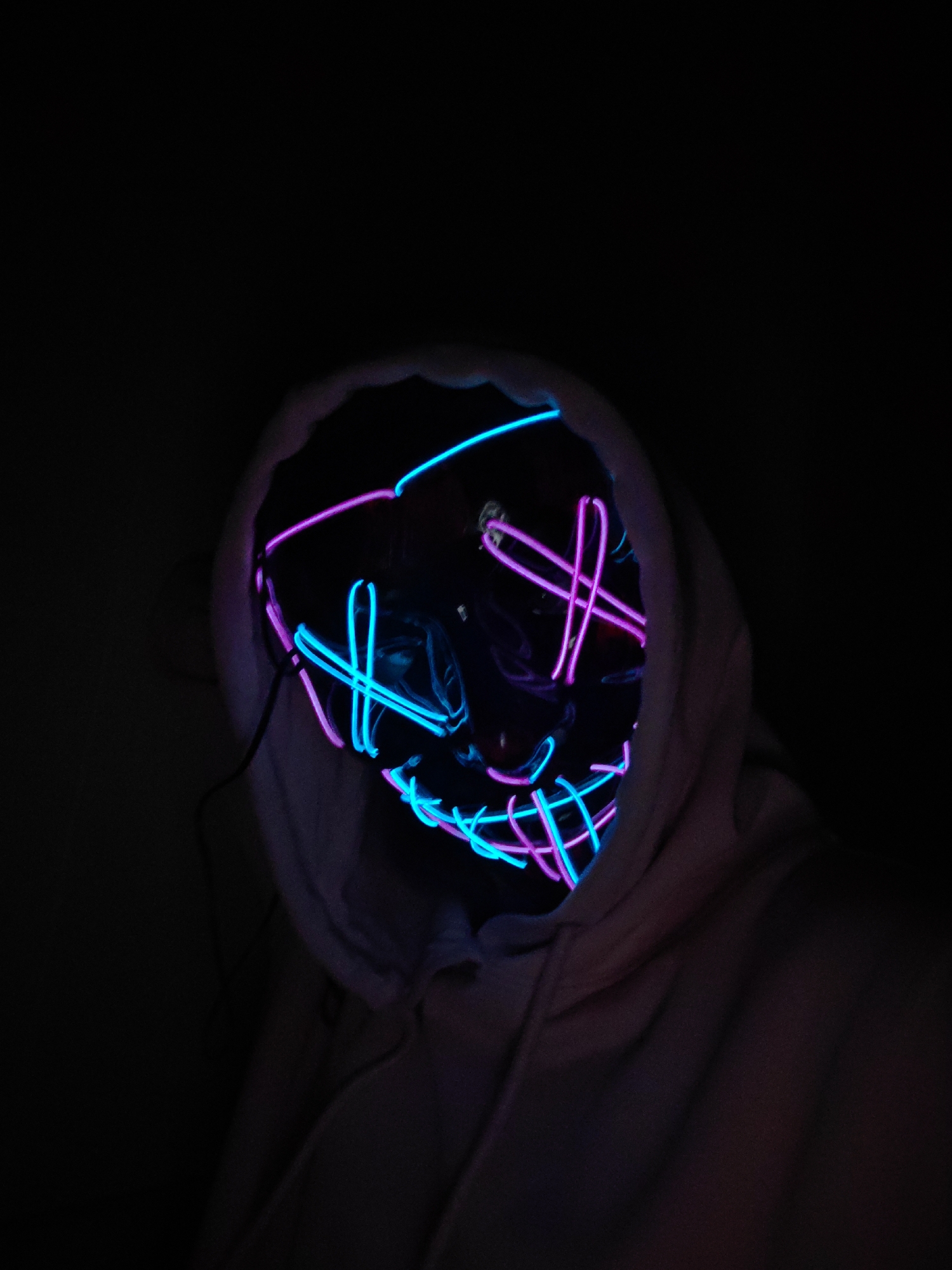 A person wearing a hooded sweatshirt and a glowing neon mask stands in a dark environment. The mask is lit with blue and pink neon lights in a geometric pattern that resembles a stylized face. The hood of the sweatshirt is pulled up, partially obscuring the person's head and shoulders. The background is black, making it difficult to discern any specific details about the environment. The photo does not contain any information about the location in Saint Petersburg, Russia.
