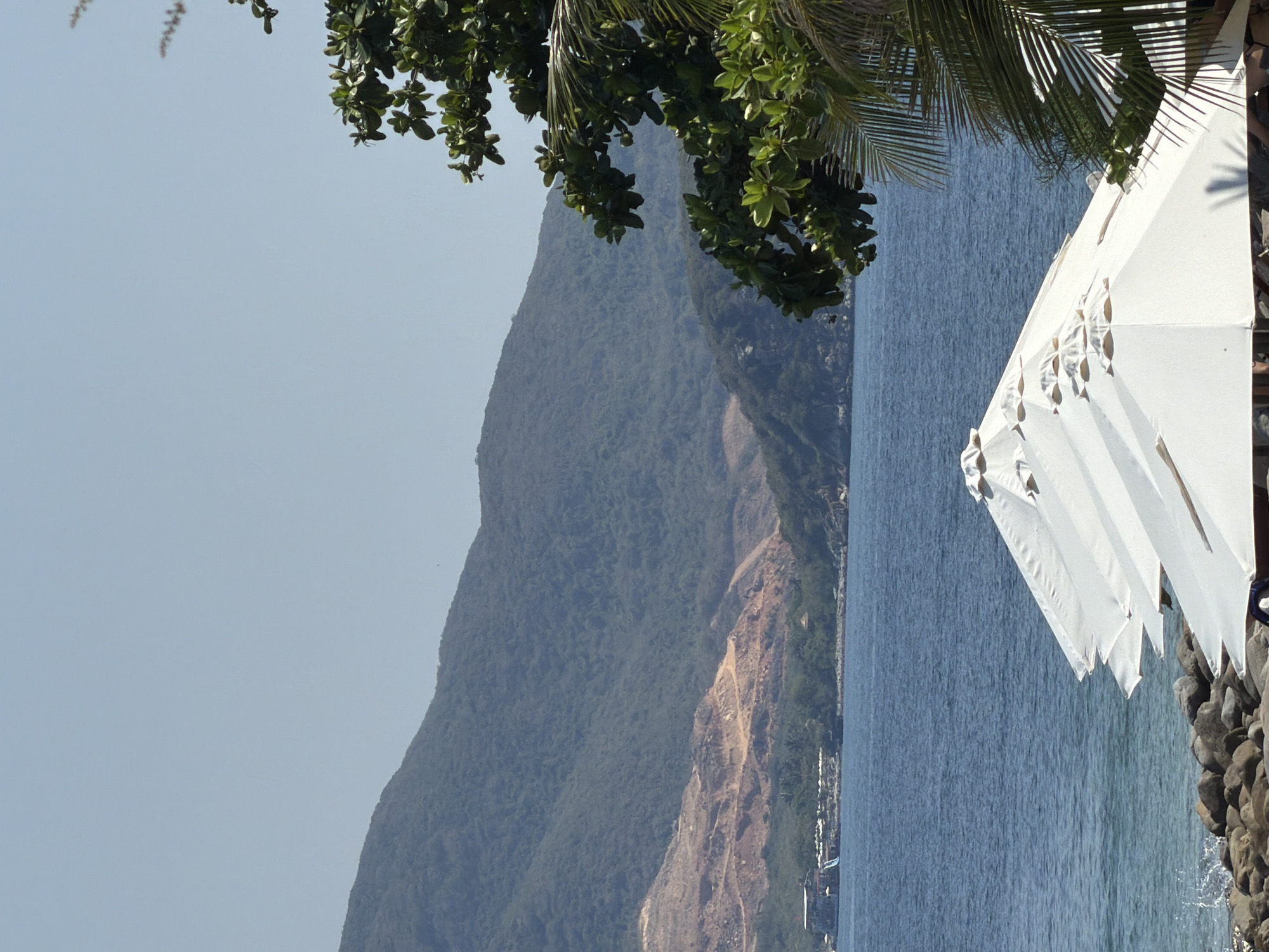 The image shows a coastal scene in Nha Trang Ward, Vietnam. In the foreground, the corner of a white canopy extends into the frame, situated on a rocky shoreline. Leaves of a tree or palm fronds hang above. The midground consists of a vast expanse of blue sea, adjacent to a large, mountainous landmass covered in dense green vegetation. A segment of the mountain reveals exposed, lighter-colored earth. The sky is clear and pale blue. Buildings and infrastructure are visible along the shoreline.
