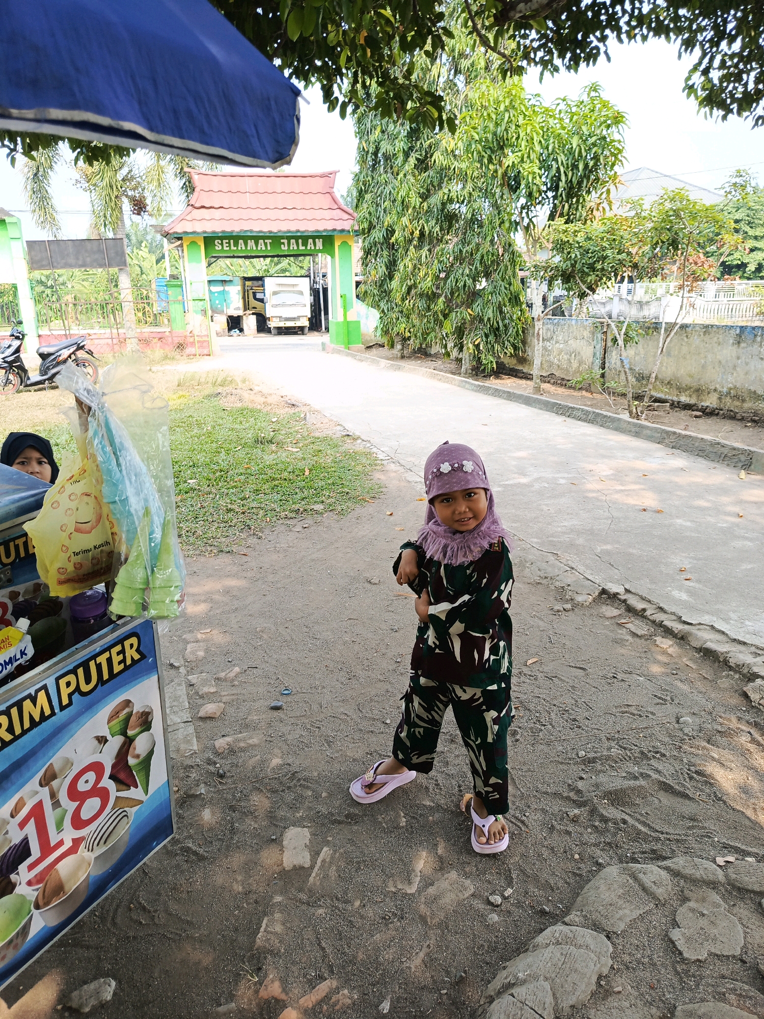 In the city of Karya Dadi, Indonesia, a young girl wearing a purple hijab and a camouflage-patterned outfit stands in front of an ice cream cart. The girl is positioned on a dirt and gravel path, with a paved road to her right. Behind her, a green and red gate with the words "SELAMAT JALAN" (which translates to "Have a good trip" or "Goodbye") is visible, indicating an exit. The gate is adorned with a red, tiled roof and is surrounded by trees and other vegetation. The ice cream cart displays images of various ice cream cones and cups, with the number "18" prominently featured. Another child, wearing a black hijab, is partially visible behind the ice cream cart. A motorcycle is parked to the left of the cart. The overall scene depicts a typical street vendor setup in a tropical environment, with the gate suggesting it might be an exit from a residential area or a checkpoint.
