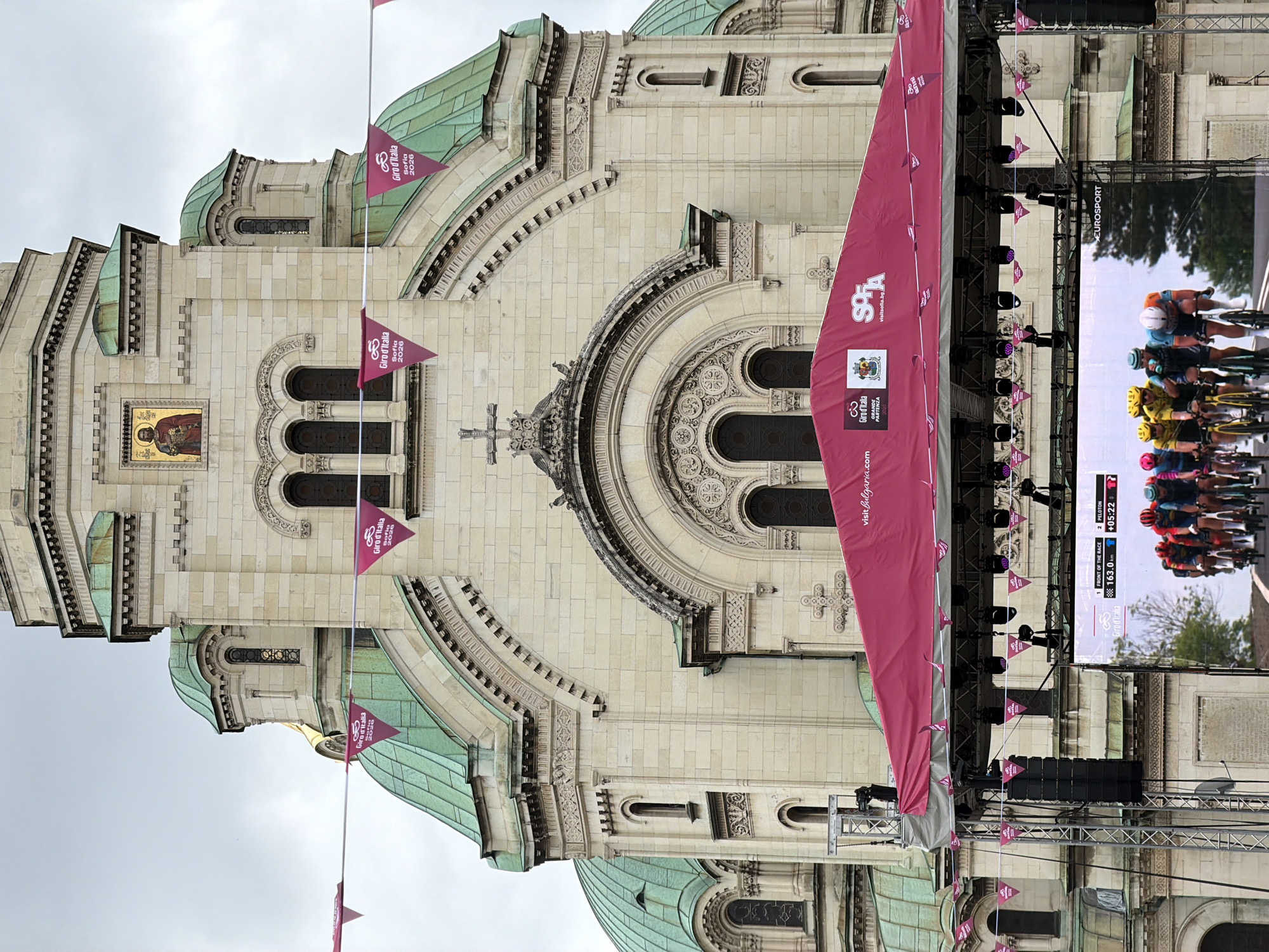 The image shows the Alexander Nevsky Cathedral in Sofia, Bulgaria. A large pink banner obscures part of the cathedral's facade. Flags with the Giro d'Italia logo are visible. A large screen displays live cycling race footage. Stage equipment, including lights and speakers, is set up in front of the building. The sky is overcast.