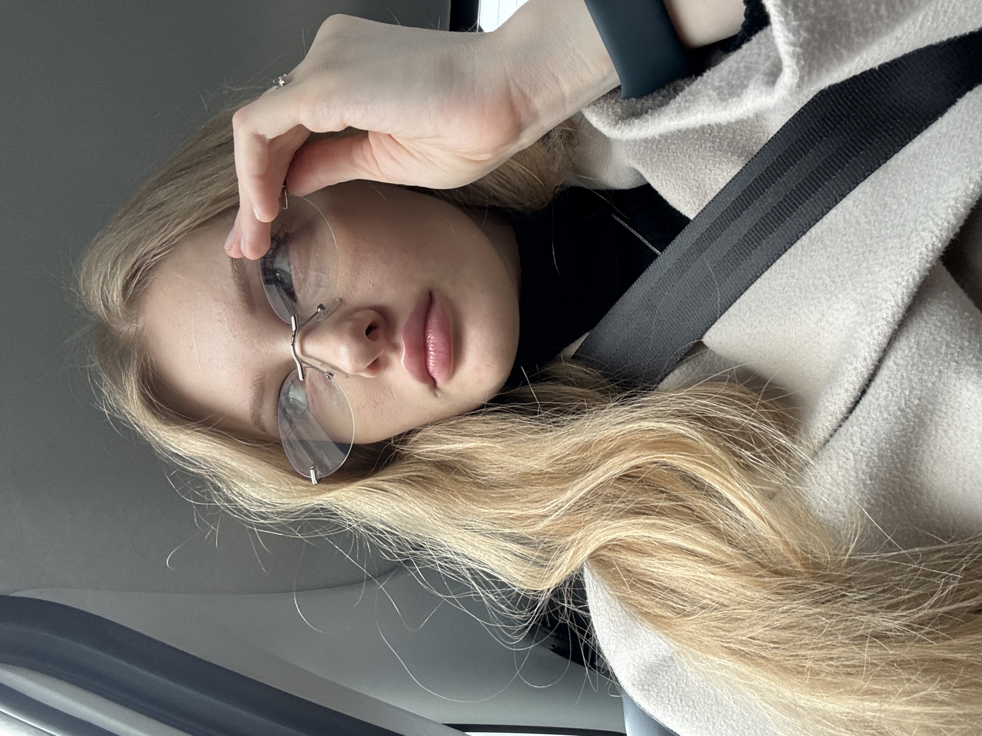 A blonde-haired woman wearing frameless glasses, a black turtleneck, and a beige coat is in a car wearing a seatbelt. Her left hand is adjusting her glasses. The photo was taken inside the car, and the outside of the car window is visible. It is difficult to determine the location in Perm, Russia.