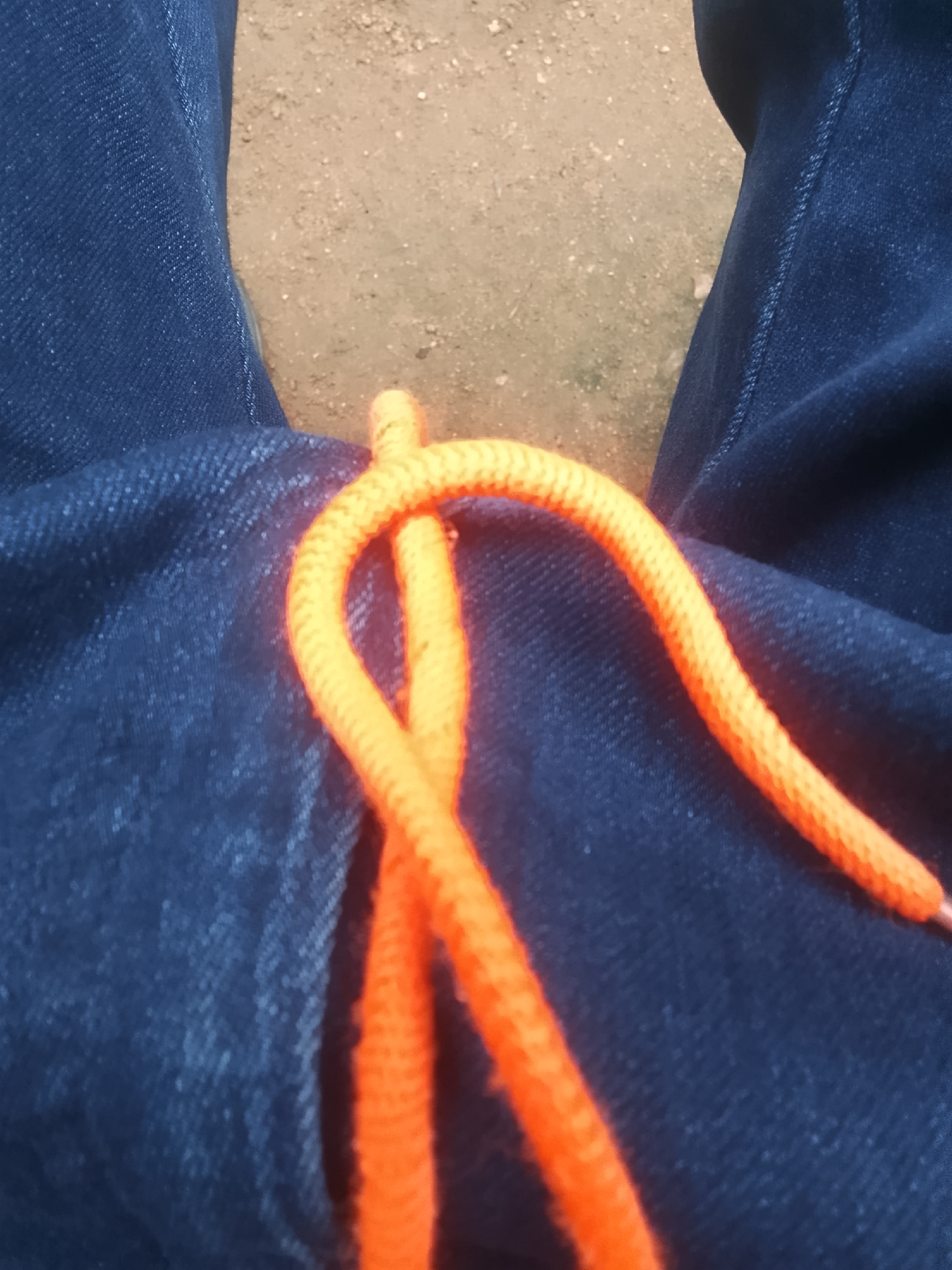 A close-up shot focuses on a pair of dark blue denim pants and an orange drawstring. The drawstring is threaded through the waistband and forms a U-shape with one end disappearing into the pants and the other end visible to the right. The background consists of a dusty, sandy surface. The scene is captured from a high angle, looking down at the pants.