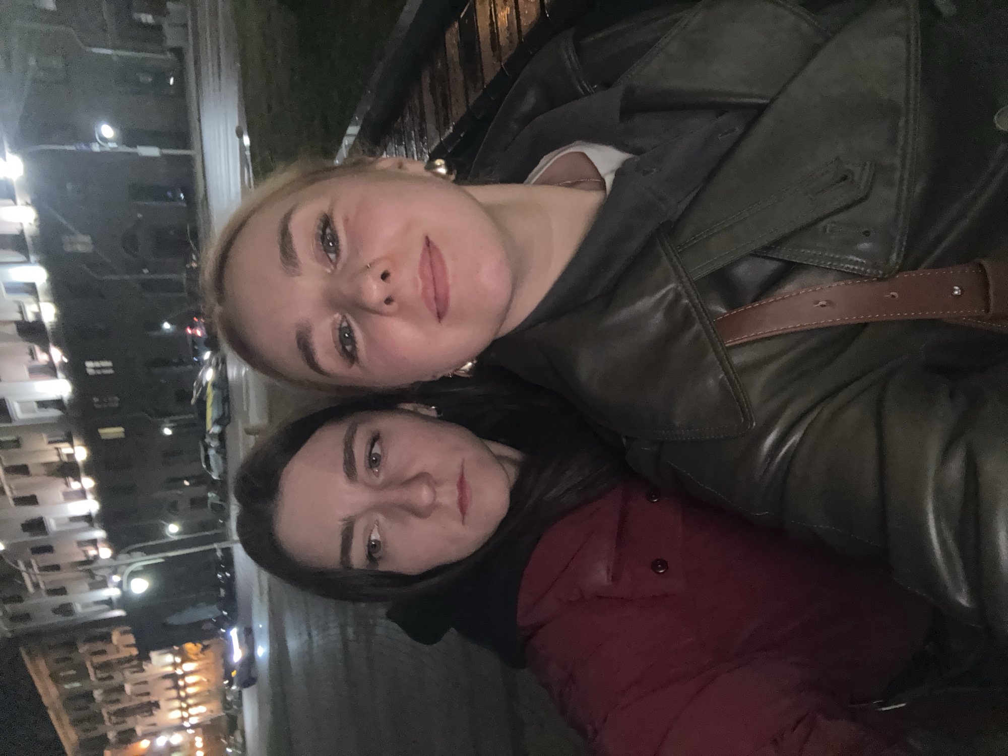 Two women are close together in an outdoor setting, likely at night, based on the lighting. The woman on the top is wearing a dark green leather jacket over a grey hoodie. A brown leather belt is visible across her chest. She has fair skin, blonde hair, and a nose piercing. The woman beneath her is wearing a red puffer jacket. She has dark hair and fair skin. The background shows buildings with many lit windows, suggesting an urban environment, possibly in Minsk, Belarus. Streetlights are also visible, contributing to the overall illumination.