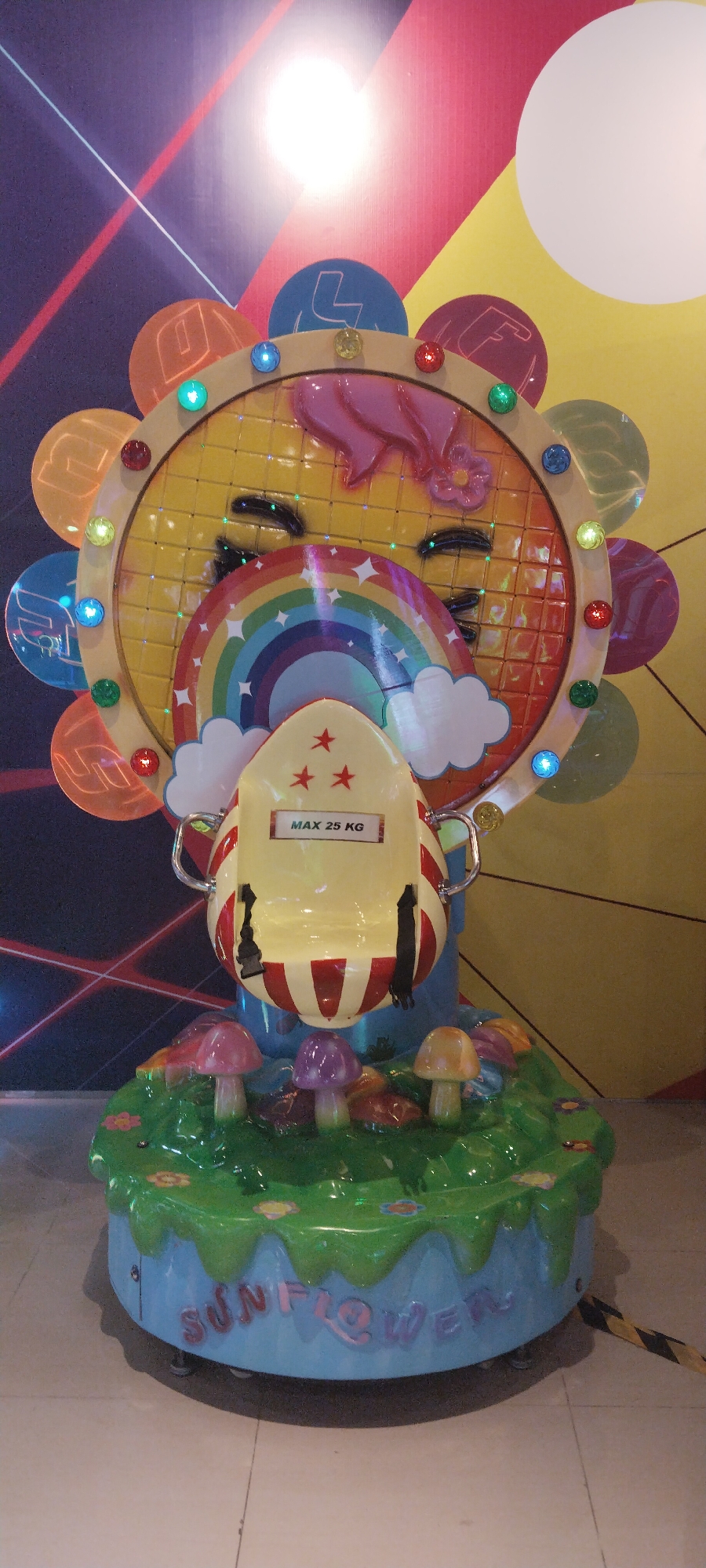 This is a medium shot, eye-level, indoor photograph of a children's amusement ride shaped like a sunflower. The ride is brightly colored with a yellow, smiling sunflower face at the center, adorned with a pink bow and a flower. A rainbow and clouds are depicted within the sunflower's face. A yellow seat with red stripes and three red stars is suspended in front of the sunflower face. A sign on the seat reads "MAX 25 KG." The ride is situated on a green, grassy base with several colorful mushrooms and small flowers. The base has the word "SUNFLOWER" written on it in raised letters. The background features a colorful, abstract pattern with diagonal lines and circles, and a bright light source. A yellow and black striped caution tape is visible in the lower right corner of the image. This amusement ride is located at the Citraland Waterpark in Semarang, Indonesia. There are no people visible in the photograph.