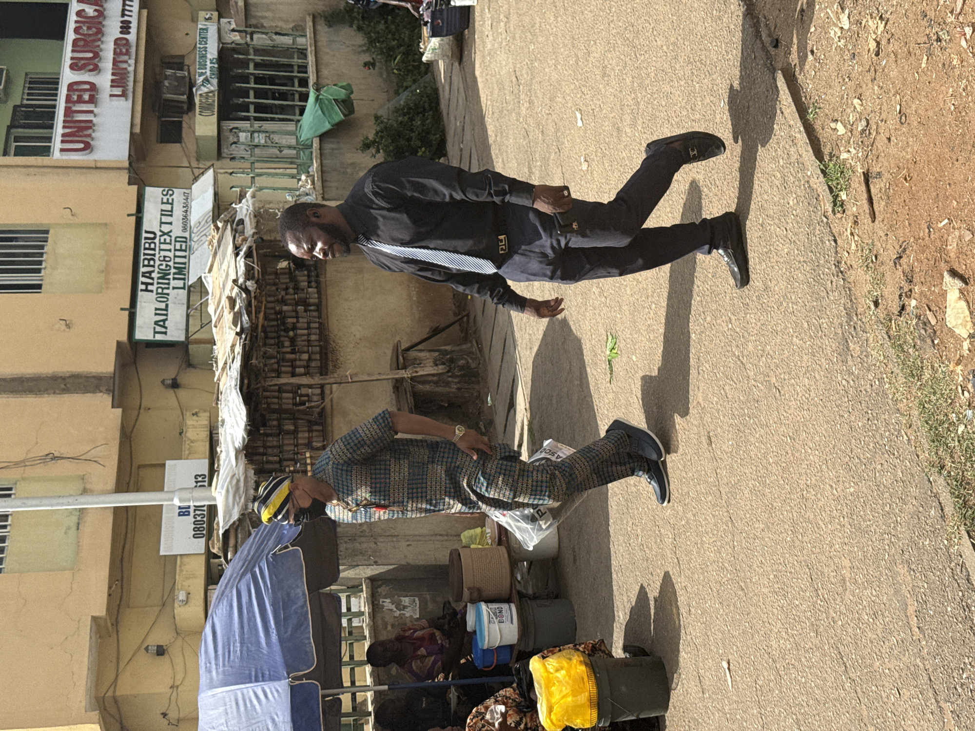 The image captures an outdoor scene in Abuja, Nigeria. Two men are walking on a paved road with signs of businesses along the left side. The first man is wearing a dark-colored suit with a striped tie. The second man is wearing a patterned top and pants, with a cap on his head. A vendor with a blue umbrella is selling goods on the side of the road. Signs for "UNITED SURGICA LIMITED", "HABIBU TAILORING & TEXTILES LIMITED", and "VE BUSINESS CENTER" are visible on the buildings. The environment suggests a commercial area in the city.