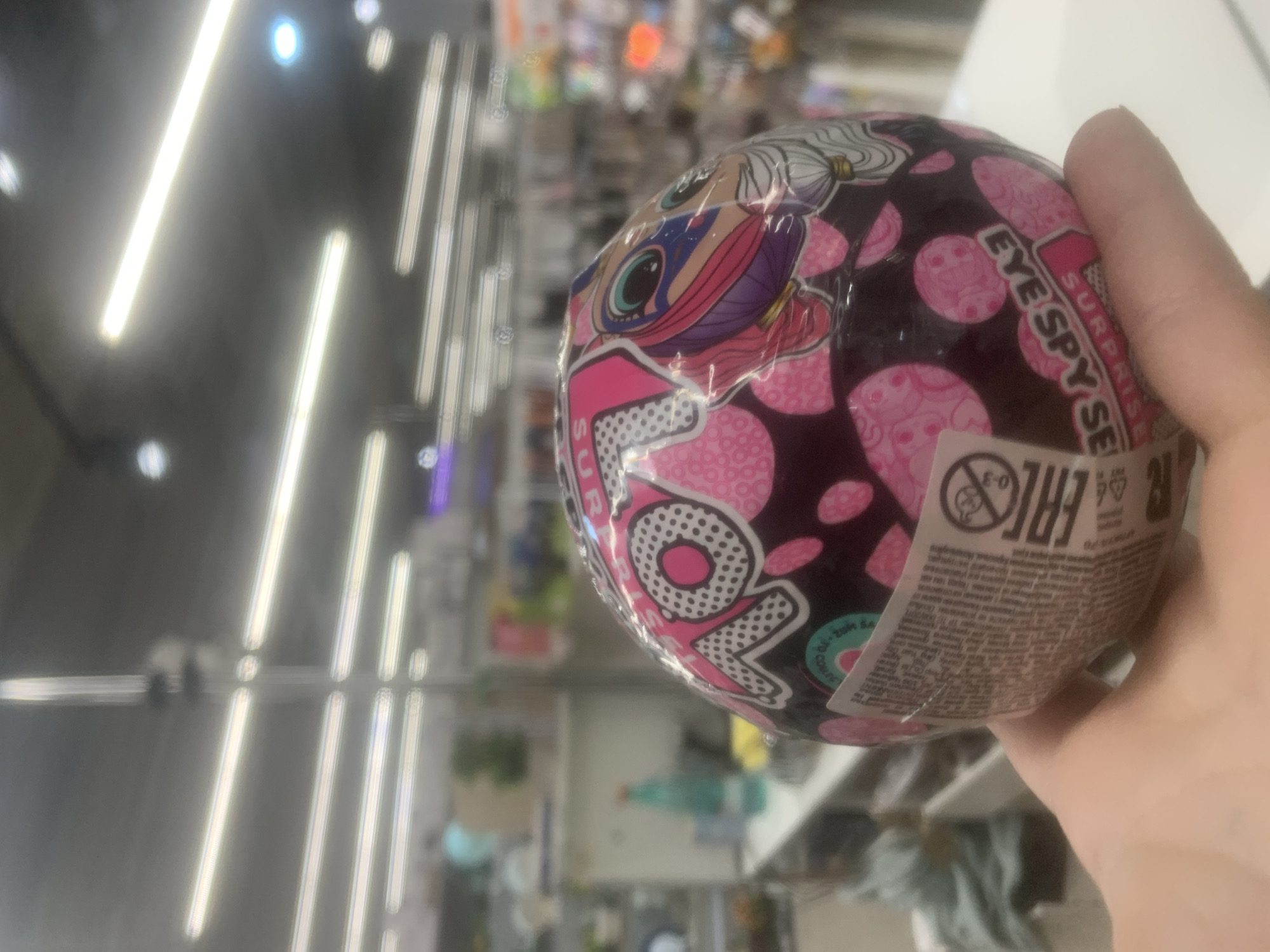 A close-up, slightly tilted image captures a person's hand holding a spherical L.O.L. Surprise! toy. The toy, predominantly black with pink polka dots, features a stylized cartoon character's face with large eyes and colorful hair. Prominently displayed text on the sphere reads "L.O.L. Surprise!" and partially visible text indicates "EYE SPY SE...", suggesting the "Eye Spy Series".
A white label affixed to the toy shows various symbols and small print. Visible symbols include the "EAC" (Eurasian Conformity) mark and a warning indicating "0-3," likely meaning not suitable for children under three years old. Some smaller, illegible text is also present on this label, likely product details or warnings.
The background is significantly blurred but reveals an indoor commercial setting, consistent with a store or supermarket aisle. Bright, elongated fluorescent lights are visible on the ceiling, creating streaks of light and reflections. Shelves stocked with various items can be faintly discerned in the depth of field. The scene takes place indoors, with artificial lighting, suggesting it could be any time of day within the commercial establishment in Saint Petersburg, Russia.