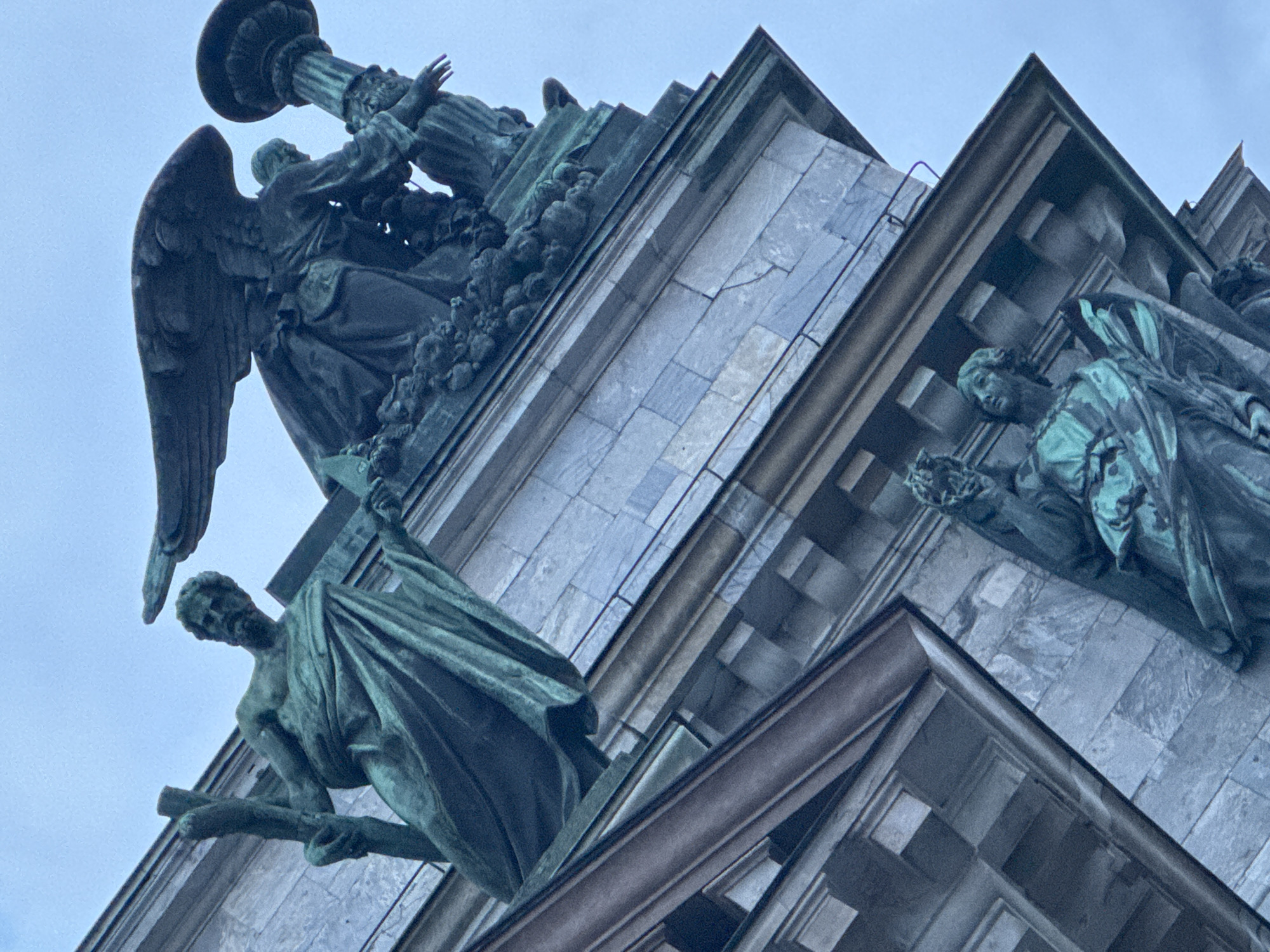 The image shows a low-angle view of a section of the Saint Isaac's Cathedral in Saint Petersburg, Russia. The architectural elements consist of stone masonry with bronze statues adorning the structure. Specifically, the statues depict religious figures, including angels, in various poses. One prominent statue features an angel with outstretched wings. The statues show green patina, indicating age and oxidation of the bronze. The background is an overcast sky.