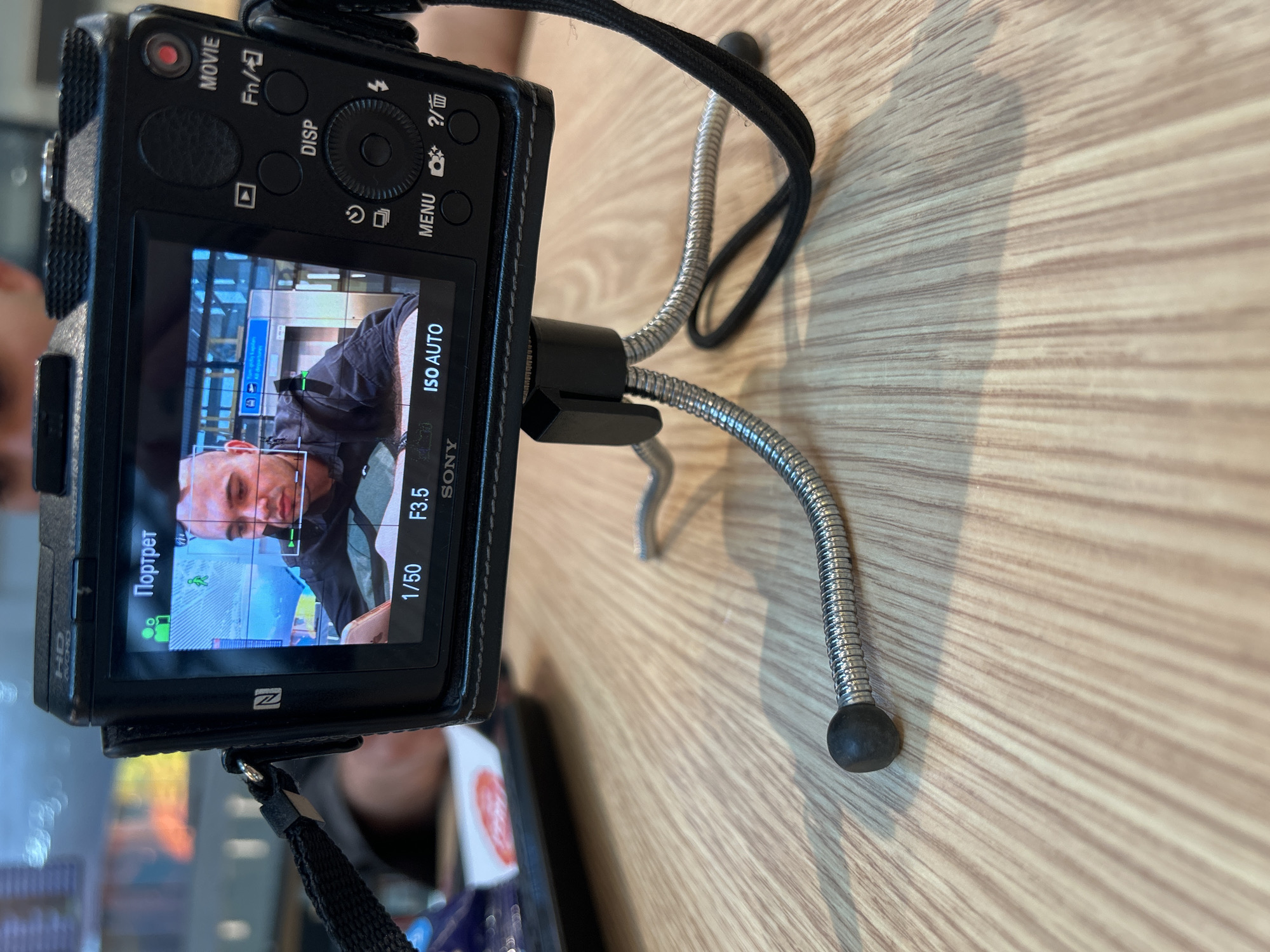 A black Sony digital camera, enclosed in a black leather-like case, is prominently displayed on a light-colored wooden table, mounted on a small, flexible tripod with silver segmented legs and black rubber feet. The camera is positioned horizontally and viewed from a slight angle.
The camera's rear LCD screen is active, showing a live view of a light-skinned man with short hair, looking directly towards the camera. He appears to be wearing a dark garment. The screen's interface displays camera settings including "Портрет" (Portrait mode), "ISO AUTO", an aperture of "F3.5", and a shutter speed of "1/50". The "SONY" logo is visible on the screen's bottom bezel, and an NFC symbol ("ن") is also present.
In the background of the live view on the camera screen, hints of an indoor public space with architectural elements and a blue sign are discernible, suggesting a location such as an airport terminal. Given the provided location context of Arnavutköy, Turkey, this setup potentially captures a scene within Istanbul Airport.
Visible buttons on the camera body include "MOVIE" (with a red indicator), "Fn/", "DISP", "MENU", and a "?/trash can" icon. The camera's textured grip is visible along its left side. The flexible legs of the tripod extend across the wooden surface, anchoring the camera in place. The overall setup indicates the camera is actively in use, likely for self-recording or photography.