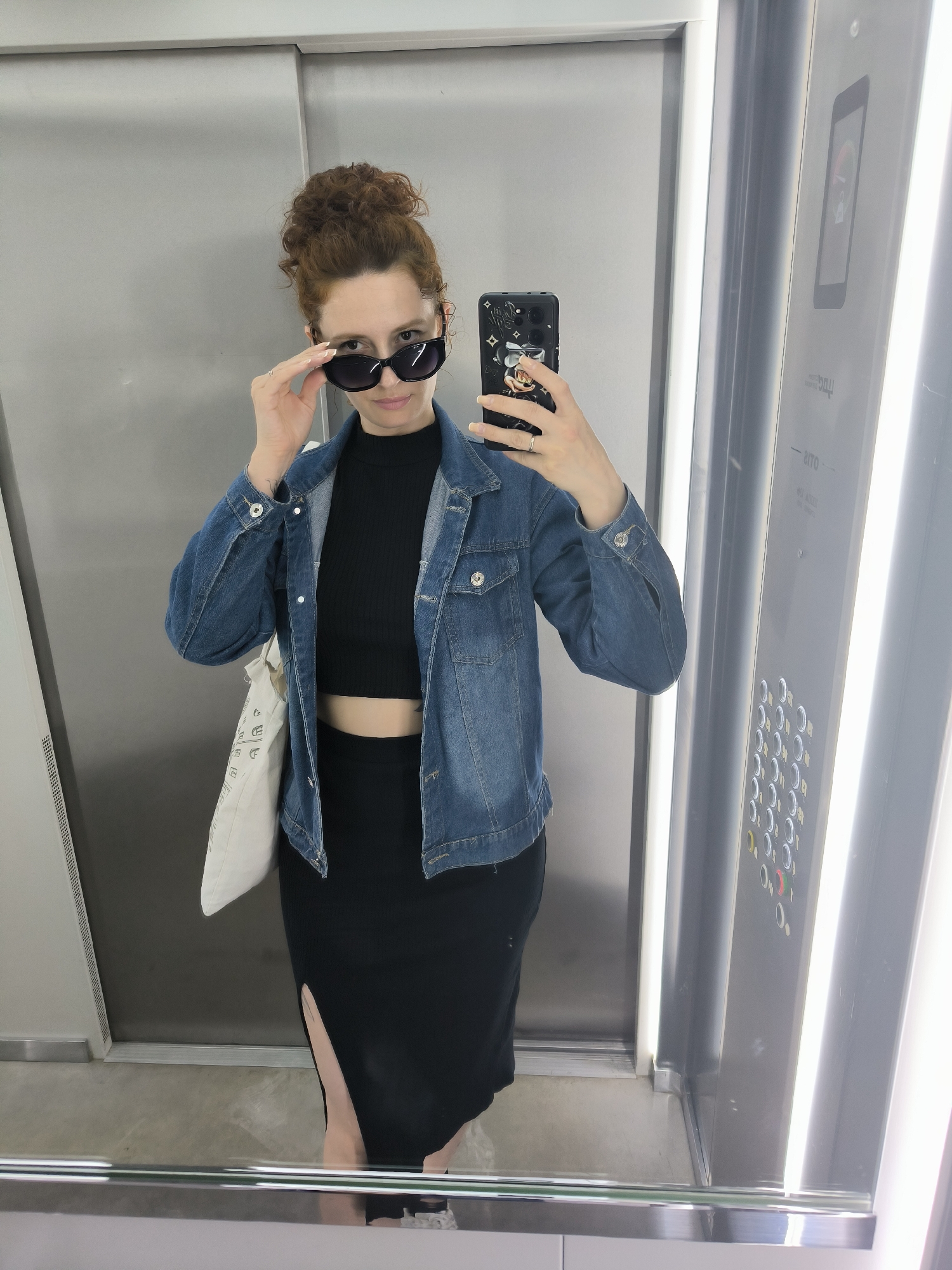 A woman with auburn hair pulled up in a messy bun is taking a selfie in an elevator. She is wearing a black crop top, a black midi skirt with a high slit, and a denim jacket. She has on large, dark sunglasses and is adjusting them with her right hand. Her left hand holds a smartphone with a Mickey Mouse case. A white tote bag with black text is slung over her left shoulder. The elevator has mirrored walls, a brushed metal finish, and bright vertical lights on the right side. A control panel with circular buttons is visible on the right wall. The floor appears to be concrete. The overall scene suggests a casual, urban setting, possibly in Saint Petersburg, Russia, during daylight hours. The woman's pose and expression are confident and playful.