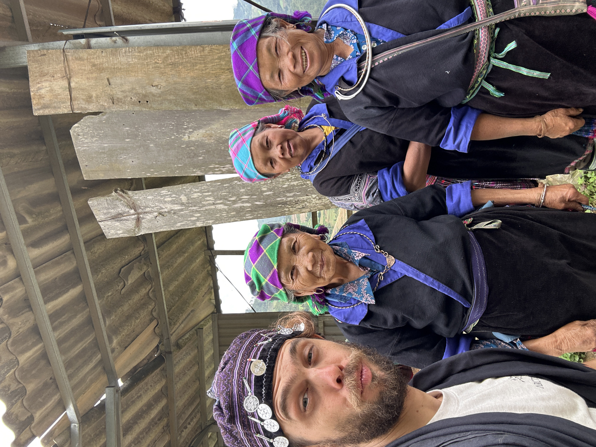 The image depicts four individuals posing for a photograph in Dế Xu Phình, Vietnam, under a corrugated fibrocement roof structure supported by metal beams.
In the foreground, on the left side, a male with a dark beard and hair looks directly at the camera with a slight pouting expression. He wears a purple and black patterned head covering embellished with silver coin-like discs, a white t-shirt with a partially visible "ARC" logo, and a dark outer garment.
Behind and to the right of the male are three women, positioned in a staggered line. They wear traditional attire, including dark, long-sleeved garments with blue accents on the collars and cuffs, and plaid headscarves. The woman closest to the male (second from left) wears a green and purple plaid headscarf and a silver necklace, exhibiting a subtle smile. The woman in the middle (third from left) has a blue and pink plaid headscarf and a silver necklace, maintaining a neutral expression towards the camera. The woman furthest to the right (fourth from left) wears a purple and blue plaid headscarf and a prominent silver collar-style necklace, smiling broadly and showing her teeth.
The background features weathered wooden support posts, some secured with wire and nails, indicating a simple, rustic construction. Beyond the structure, a hazy, green mountainous landscape is visible under an overcast sky, consistent with the rural, mountainous terrain of Dế Xu Phình. Sparse green vegetation is visible at the bottom of the frame.