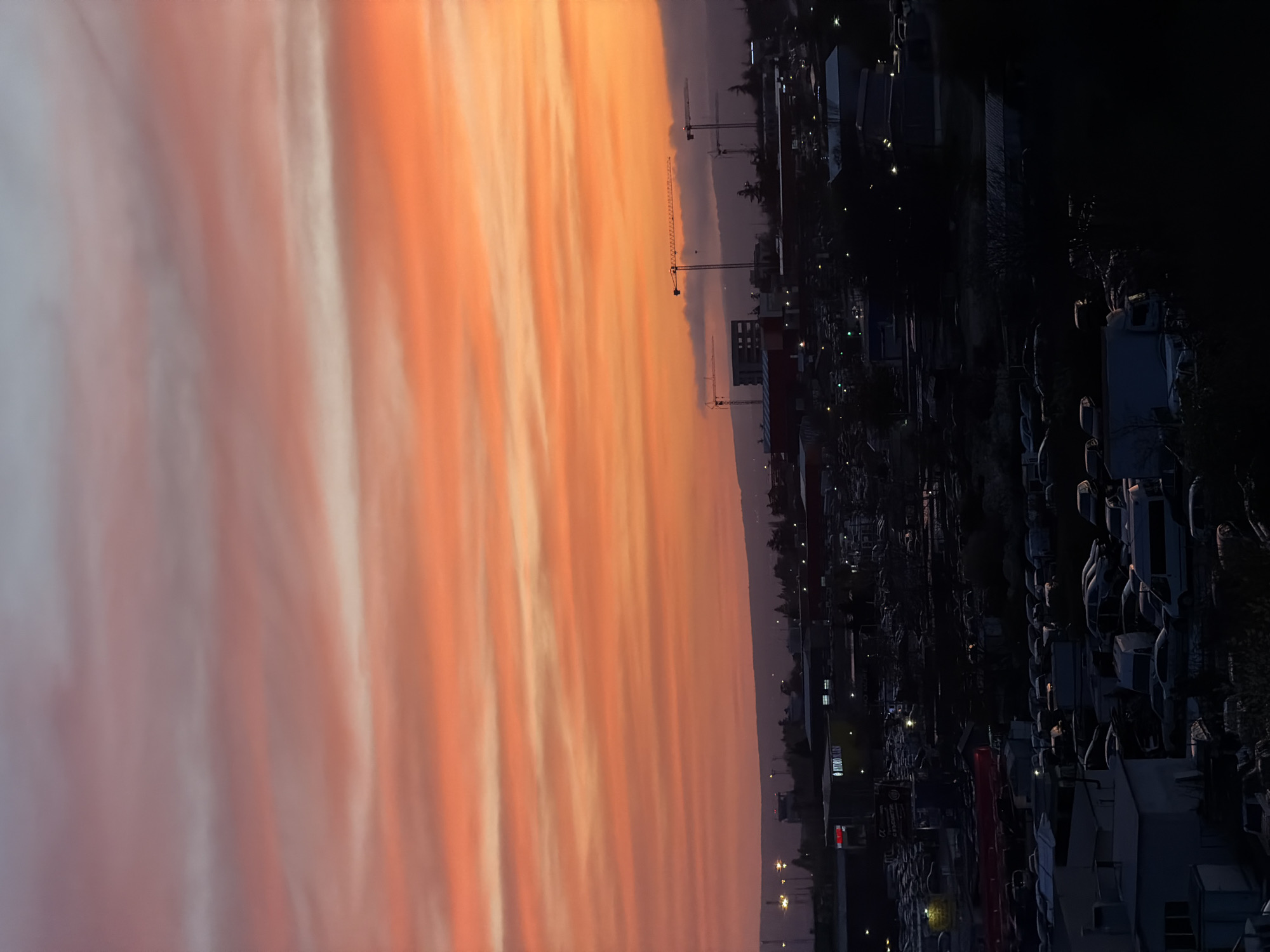 The image captures a vibrant sunset or sunrise over an urban or industrial landscape in Varna, Bulgaria. The sky dominates the upper portion of the frame, painted with dramatic, elongated clouds in brilliant shades of orange, red, and pink, transitioning to softer white and grey hues towards the left. The intensity of the colors suggests the golden hour of dusk or dawn.
Against this colorful backdrop, the silhouette of a city or industrial area stretches along the horizon. Several tall construction cranes are prominently visible, indicating ongoing development. One particularly large crane stands out against the glowing sky. Various buildings are present, mostly as dark shapes, but some show subtle outlines or faint internal lights.
The lower half of the image is much darker, revealing a complex array of structures and vehicles. A large area appears to be a parking lot or storage yard, densely packed with numerous cars, vans, or possibly boats on trailers, reflecting the context of Varna as a port city. Scattered artificial lights from buildings and streetlights punctuate the darkness, adding small points of illumination to the otherwise shadowy foreground.
Visible text includes the word "HYUNDAI" in white letters on a building or sign, and possibly "MOTORSPORT" or a similar phrase below it, though less clearly discernible. No people or specific activities are visible, emphasizing the expansive landscape and the striking natural phenomenon of the colorful sky. The overall mood is one of serene contemplation, highlighting the dramatic interplay of natural light and urban structures.