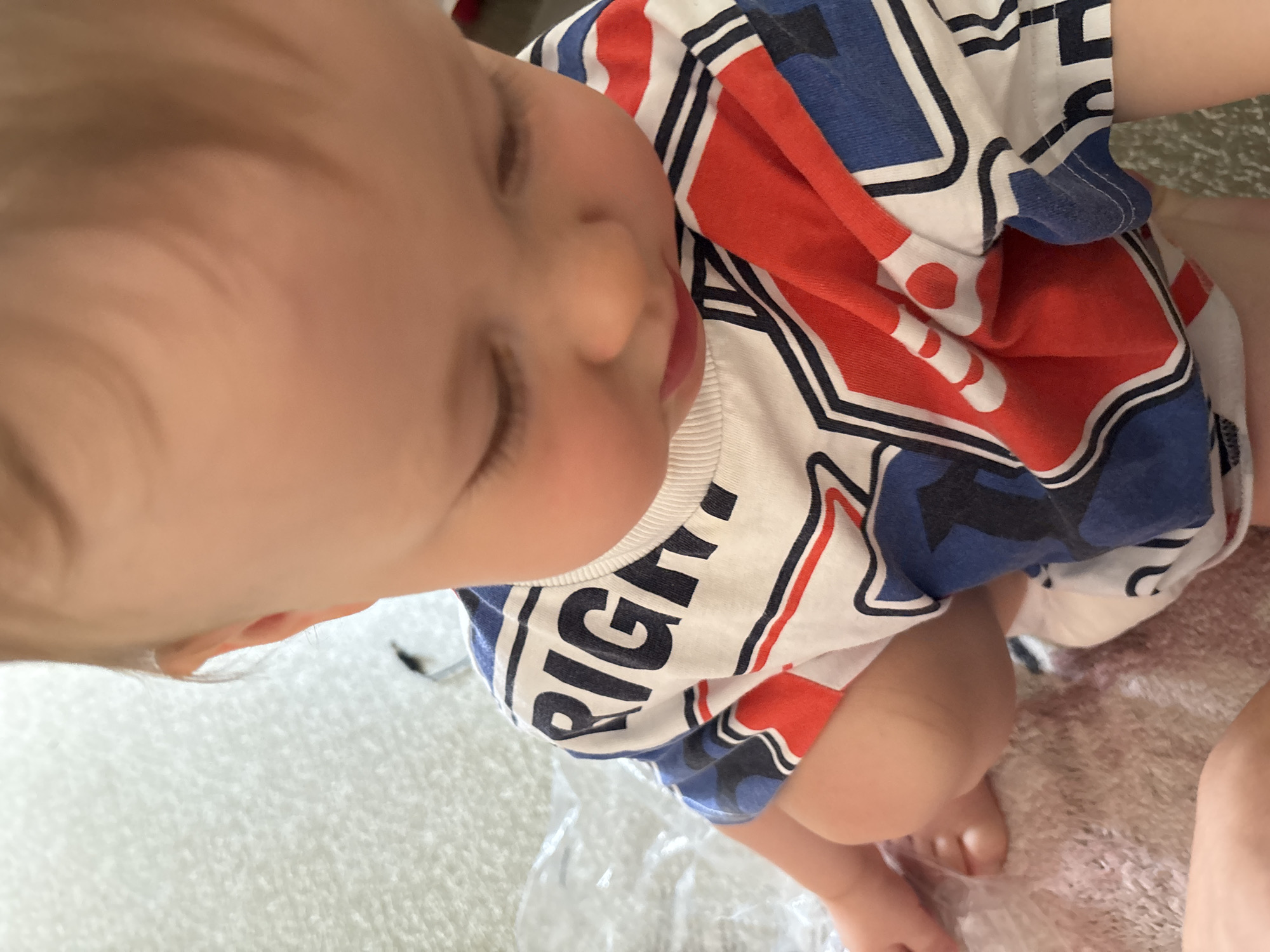 The image shows a close-up, top-down view of a baby wearing a t-shirt with a red, white, and blue pattern. The baby is looking down, with their head tilted forward. The t-shirt has the word "RIGHT" in large black letters across the chest. The background is blurred and appears to be a textured surface, possibly a carpet or rug. There are no identifiable landmarks or elements in the image that would indicate a specific location within Naberezhnye Chelny, Russia. The image focuses solely on the baby and their clothing.