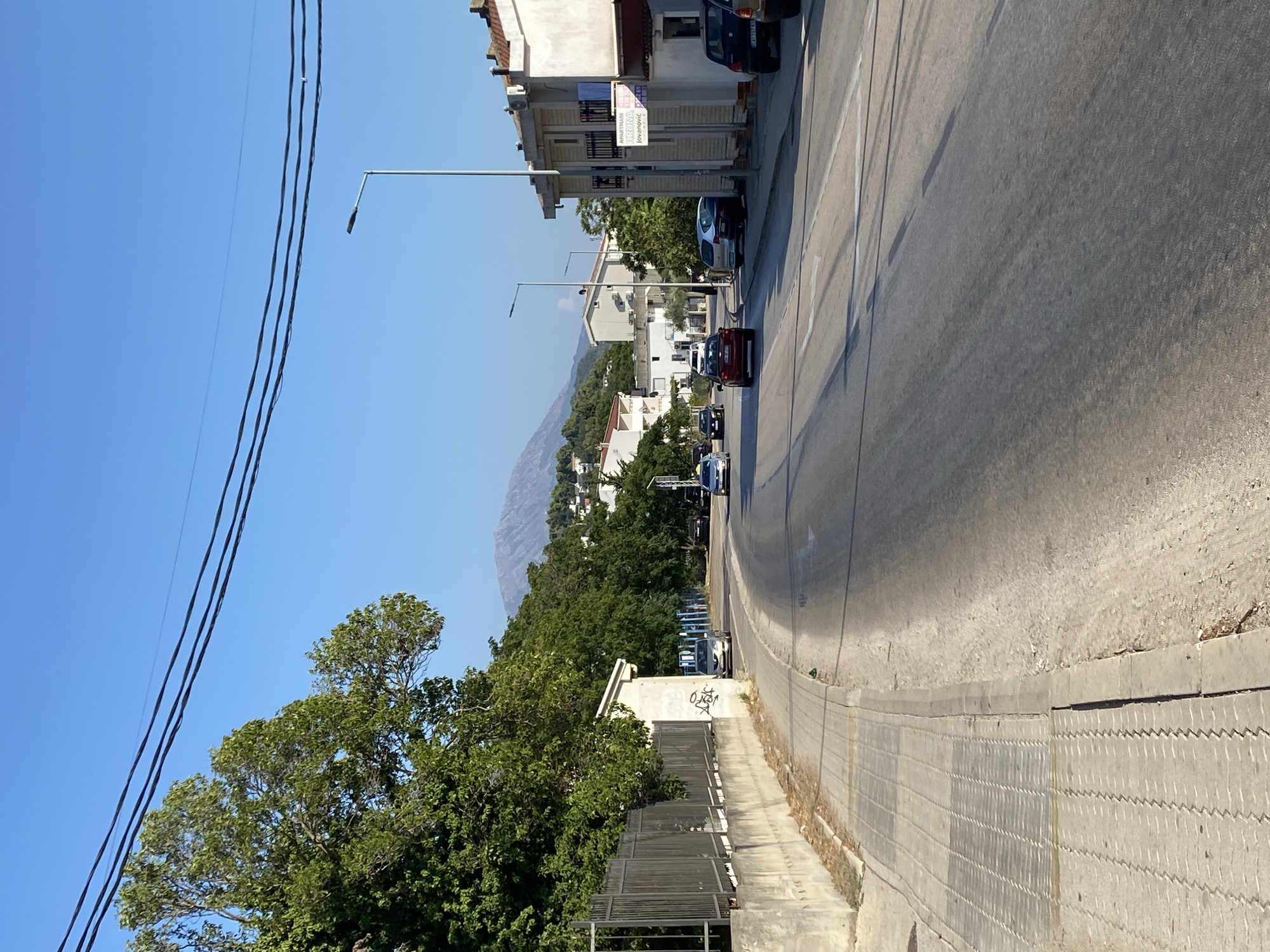 The image depicts a street scene in Sutomore, Montenegro, on a clear, sunny day. The perspective is from the side of the road, looking down a paved street that curves to the left. On the left side of the street, there is a sidewalk made of textured concrete tiles, bordered by a low concrete wall and a metal fence. Lush green trees line the left side of the road beyond the fence.
The road itself is asphalt and appears to be a two-lane road with visible lane markings, including arrows. Several cars are present on the road. On the right side of the street, there are multi-story buildings with white and beige facades, some featuring balconies. One building has a sign with text that appears to be in a Slavic language, indicating "Apartmani Jovanovic". Streetlights are visible along the right side of the road.
In the distance, a large, hazy mountain range dominates the skyline under a bright blue sky. Overhead, several dark power lines stretch across the frame. The overall impression is of a moderately busy street in a coastal town, with elements of urban development set against a natural, mountainous backdrop.