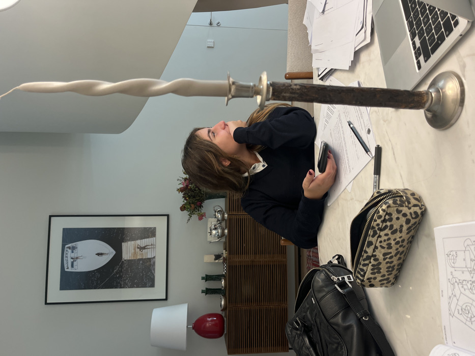 An adult female is seated at a light-colored table in an interior space within Lordelo do Ouro e Massarelos, Portugal. The woman, with long brown hair, wears a dark blue V-neck sweater over a white collared shirt. Her head is turned to the right, and her right hand rests on her chin. Her left hand holds a dark-colored smartphone. On the table to her left, a black leather bag and an open leopard-print pencil case are present. Directly in front of her, white papers with text and diagrams, along with a black pen and marker, are visible. A silver-colored laptop is partially visible to her right. In the foreground, a tall, dark-colored candlestick with a silver base and top holds a light-grey twisted candle. The background features a light-colored wall with a large framed black-and-white print depicting a boat and people on water and a pier. Beneath the print, a wooden slatted sideboard holds a red ceramic lamp with a white shade, two green decorative objects, a white sculpture, a silver tea set, and a vase of flowers. To the far right, a segment of a white modern staircase and a light-colored door with a metallic handle are visible.