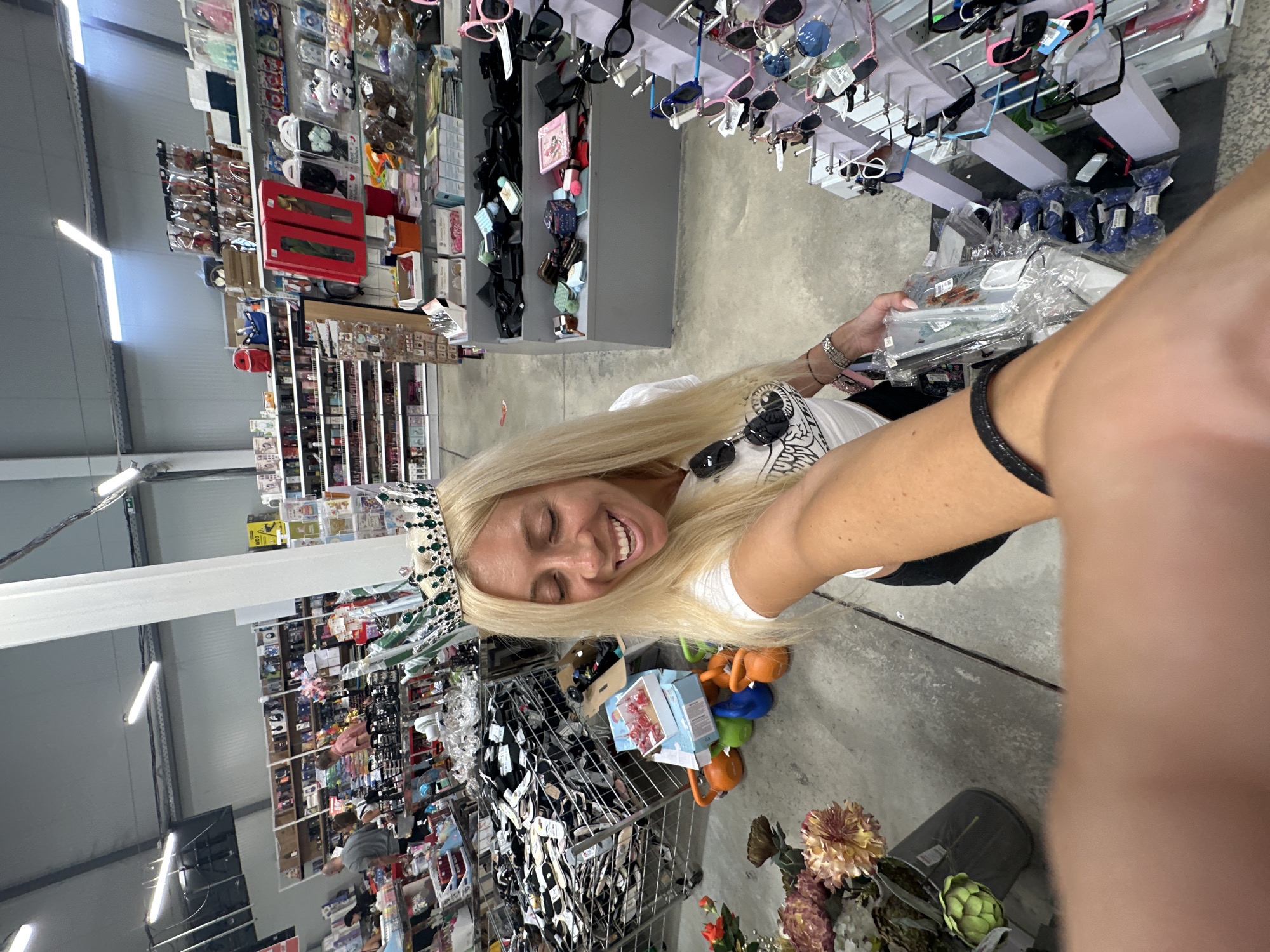 A blonde woman wearing a crown and a white t-shirt is standing in what appears to be a large retail store or warehouse. She is smiling and holding a clear plastic package in her right hand. The store is stocked with shelves of merchandise, including clothing, accessories, and other retail items. In the background, other shoppers can be seen browsing the aisles. The flooring is concrete, and fluorescent lighting illuminates the space. The overall environment suggests a busy commercial setting. The image does not provide specific details to determine the city of Pazardzhik, Bulgaria.
